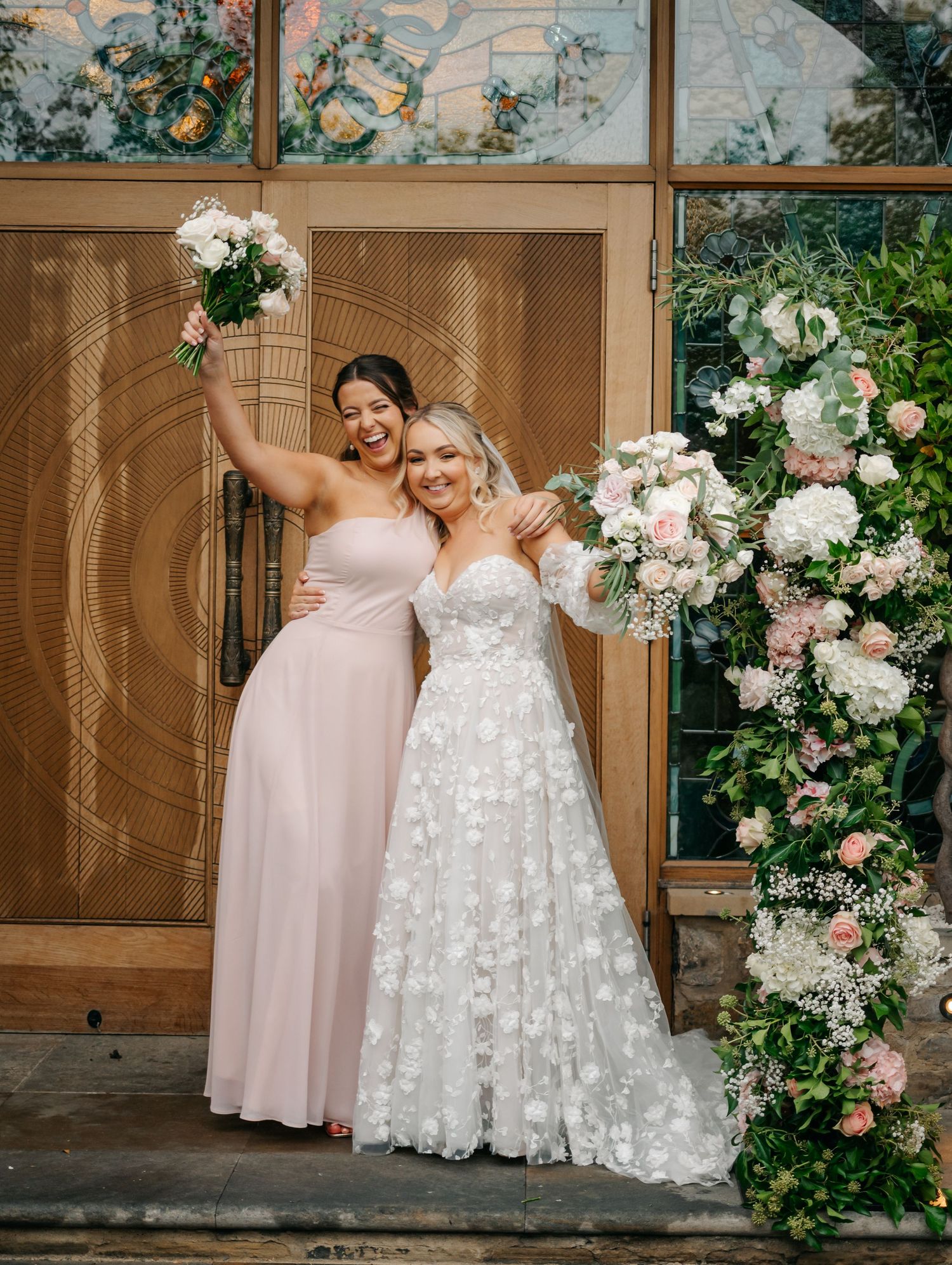 Elegant wedding party poses against a floral arch with white and pink roses in front of wooden doors.