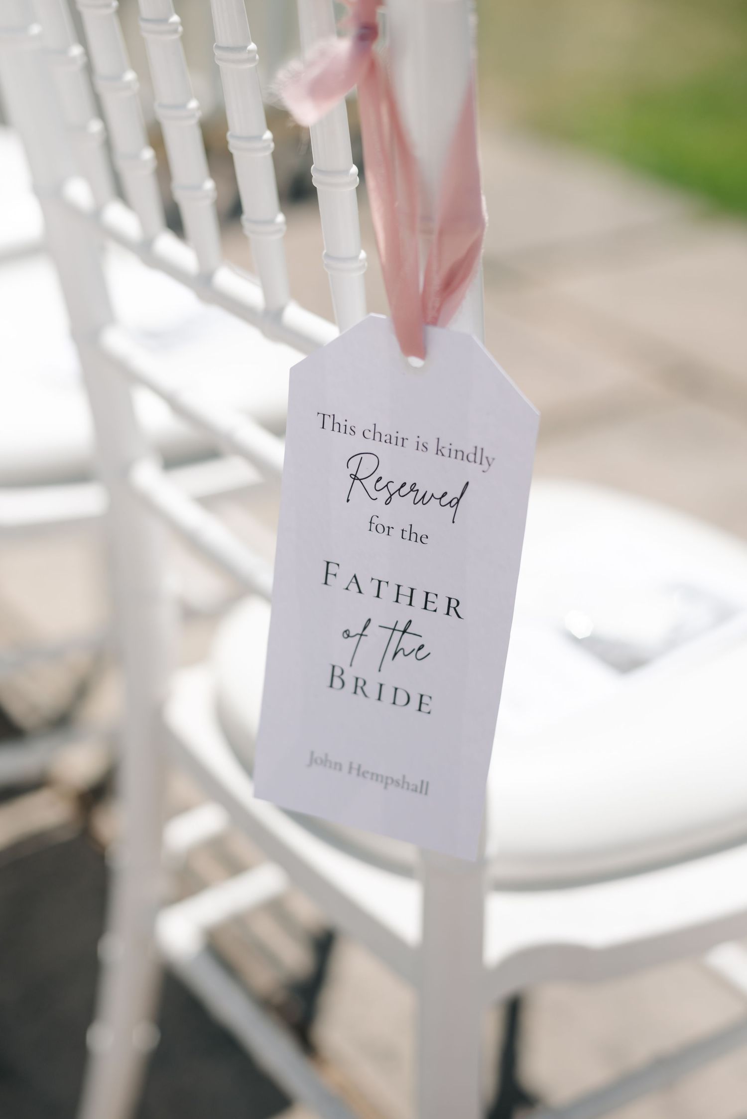 Elegant white chair with reserved sign for Father of the Bride tied with pink ribbon at outdoor wedding ceremony.