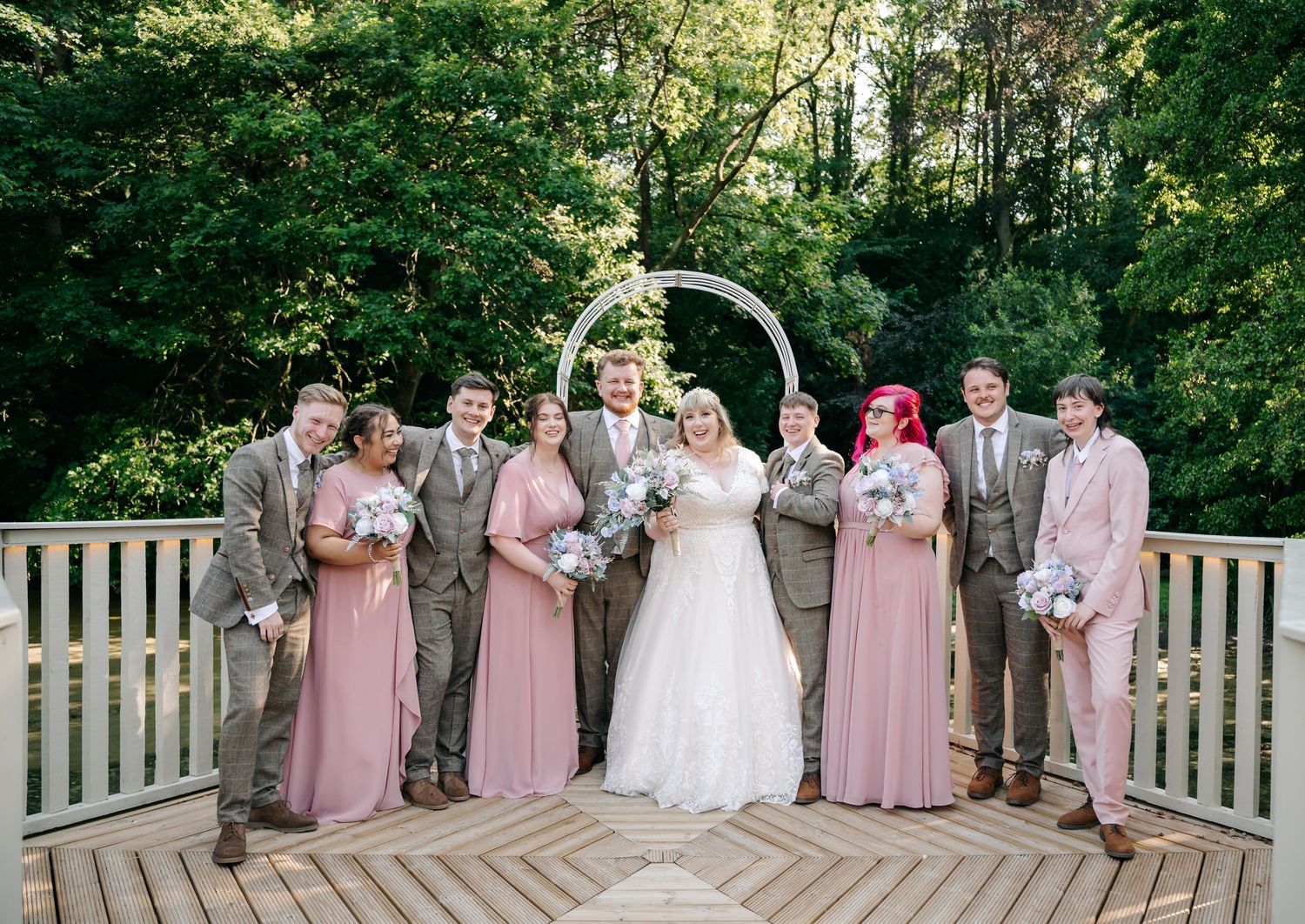 Wedding party poses on wooden deck with circular arch, bridesmaids in pink gowns and groomsmen in tan suits.