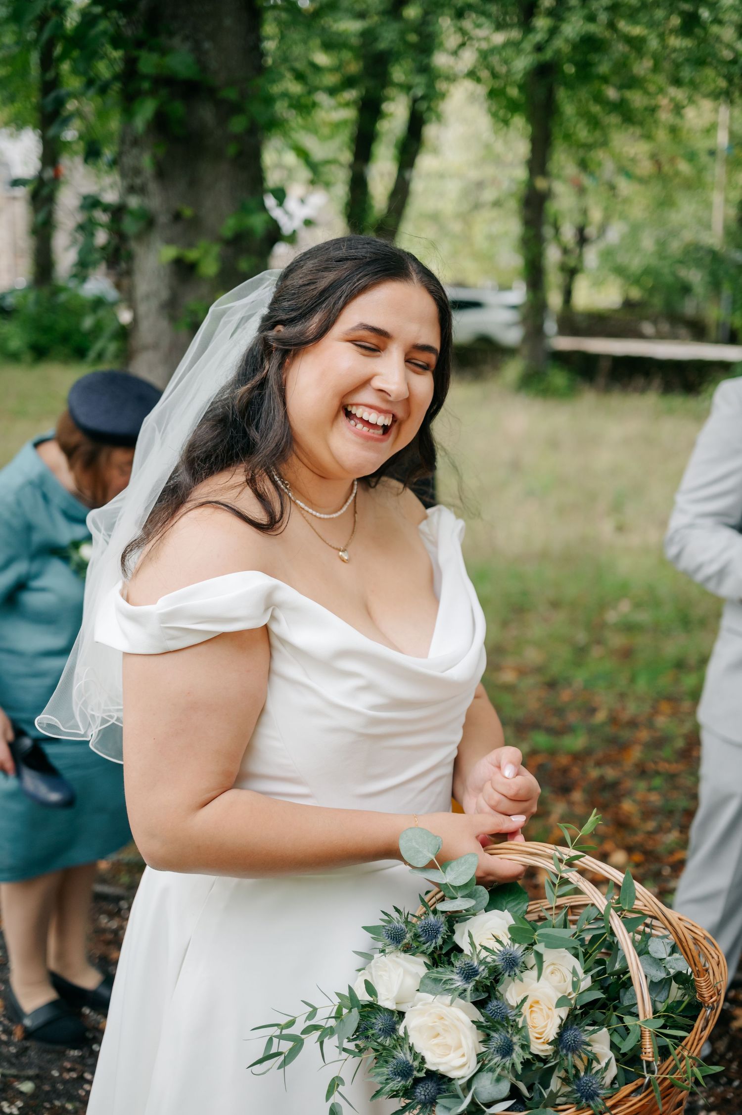 Bride in white dress holds a floral basket while smiling joyfully outdoors.