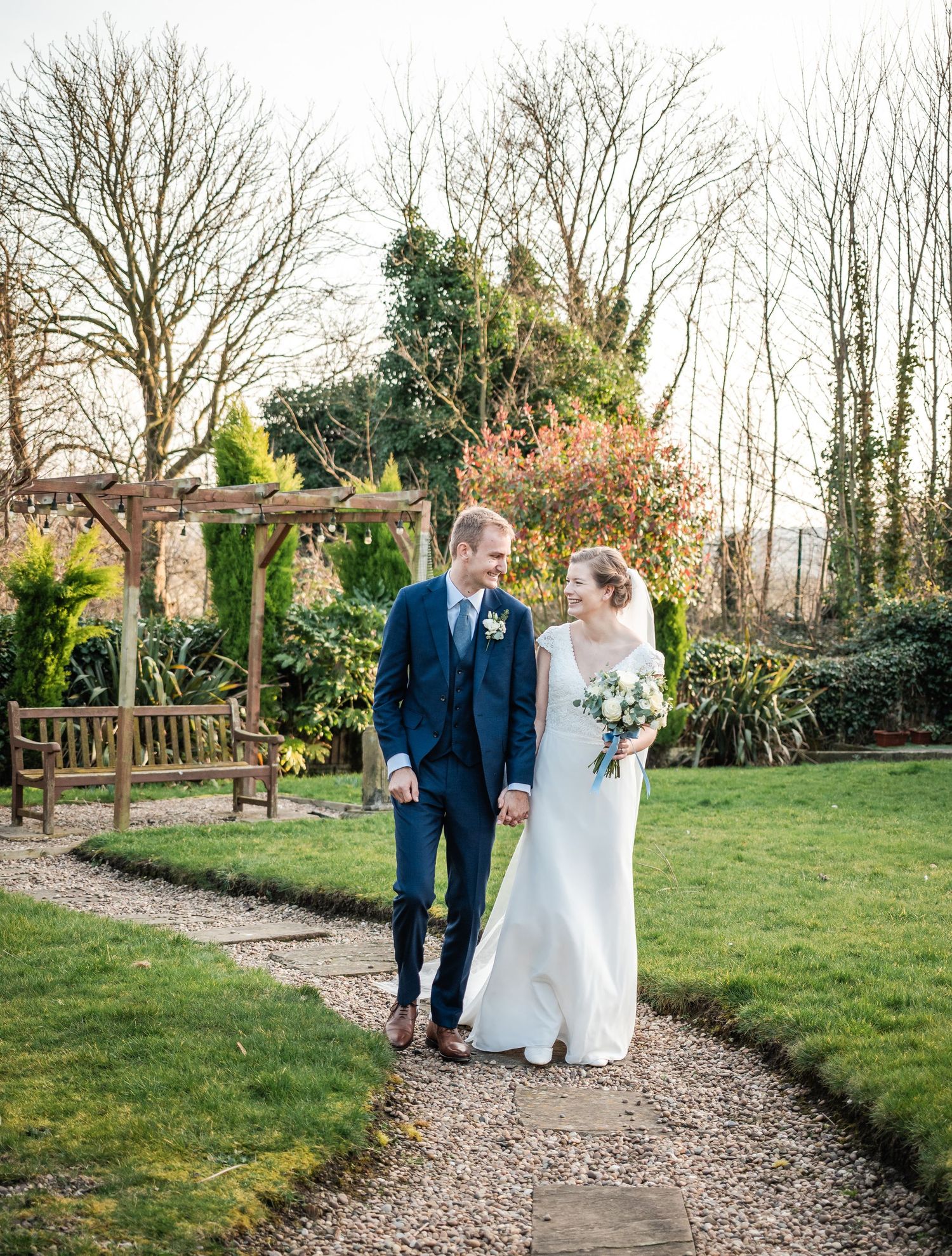 Wedding couple walking along a garden path surrounded by winter trees and greenery.