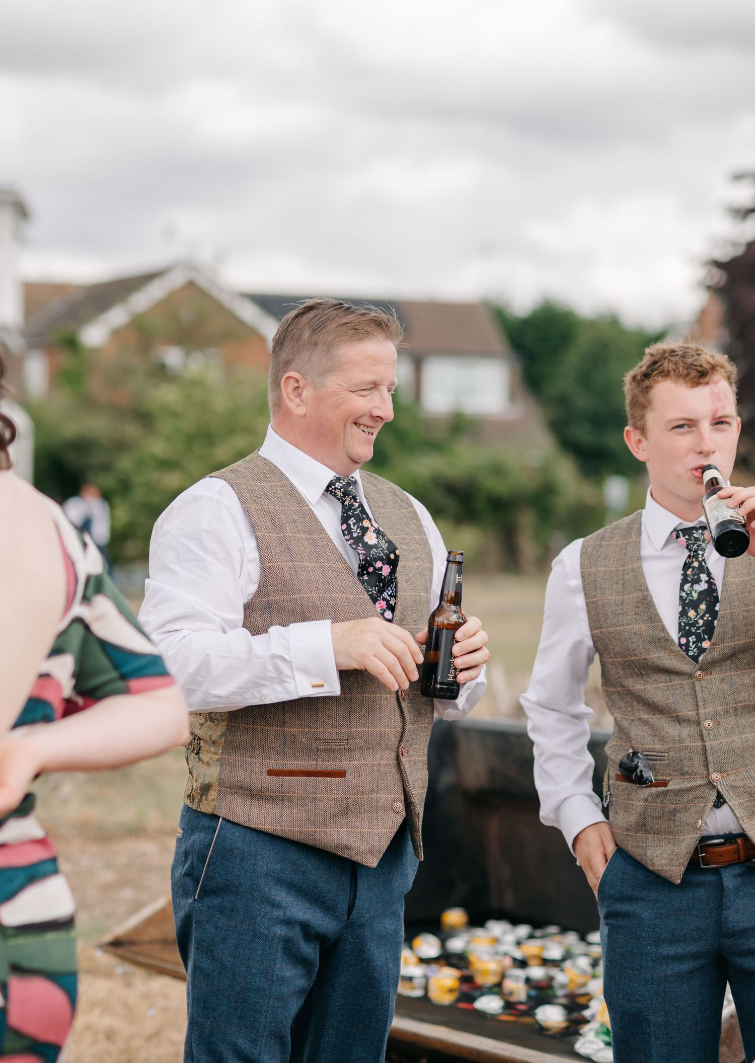 Wedding guests in vests and ties enjoying drinks outdoors.