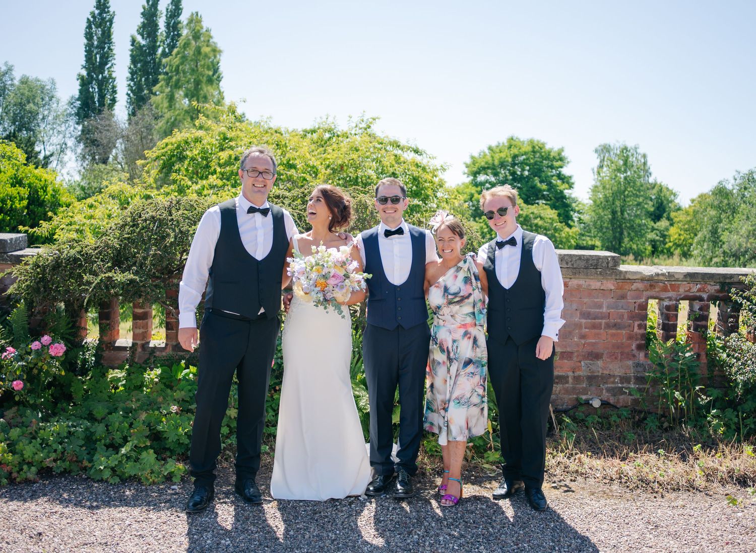 Wedding party members walk and pose together on a gravel path surrounded by lush garden landscaping.