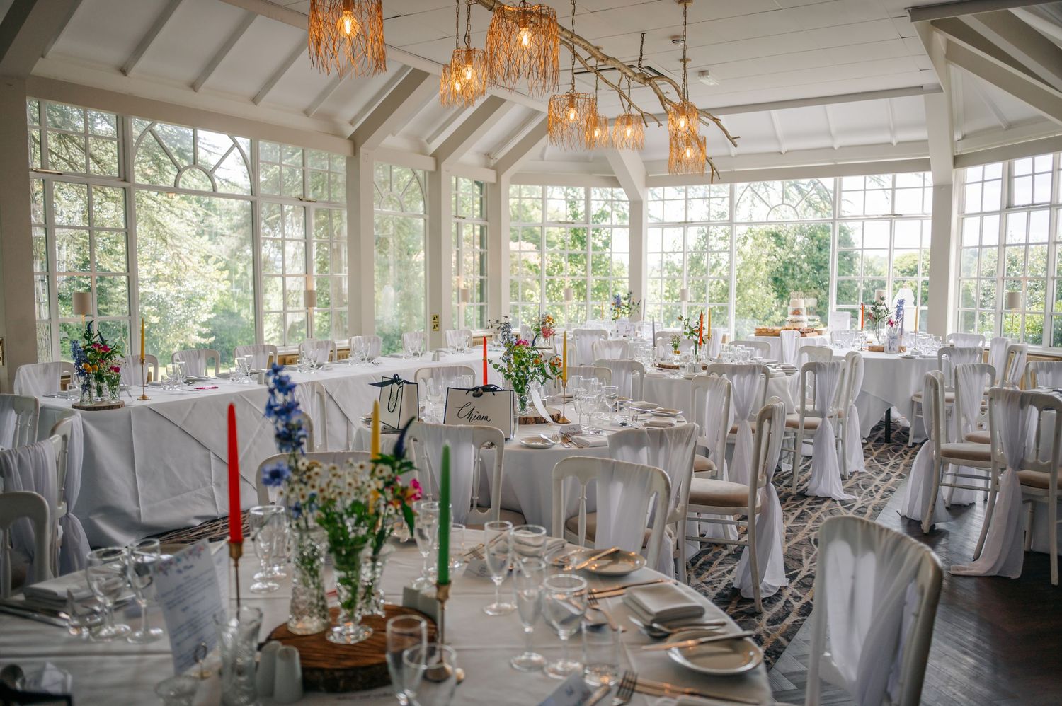 An elegant wedding reception venue with white decor, chandeliers and large windows overlooking gardens.