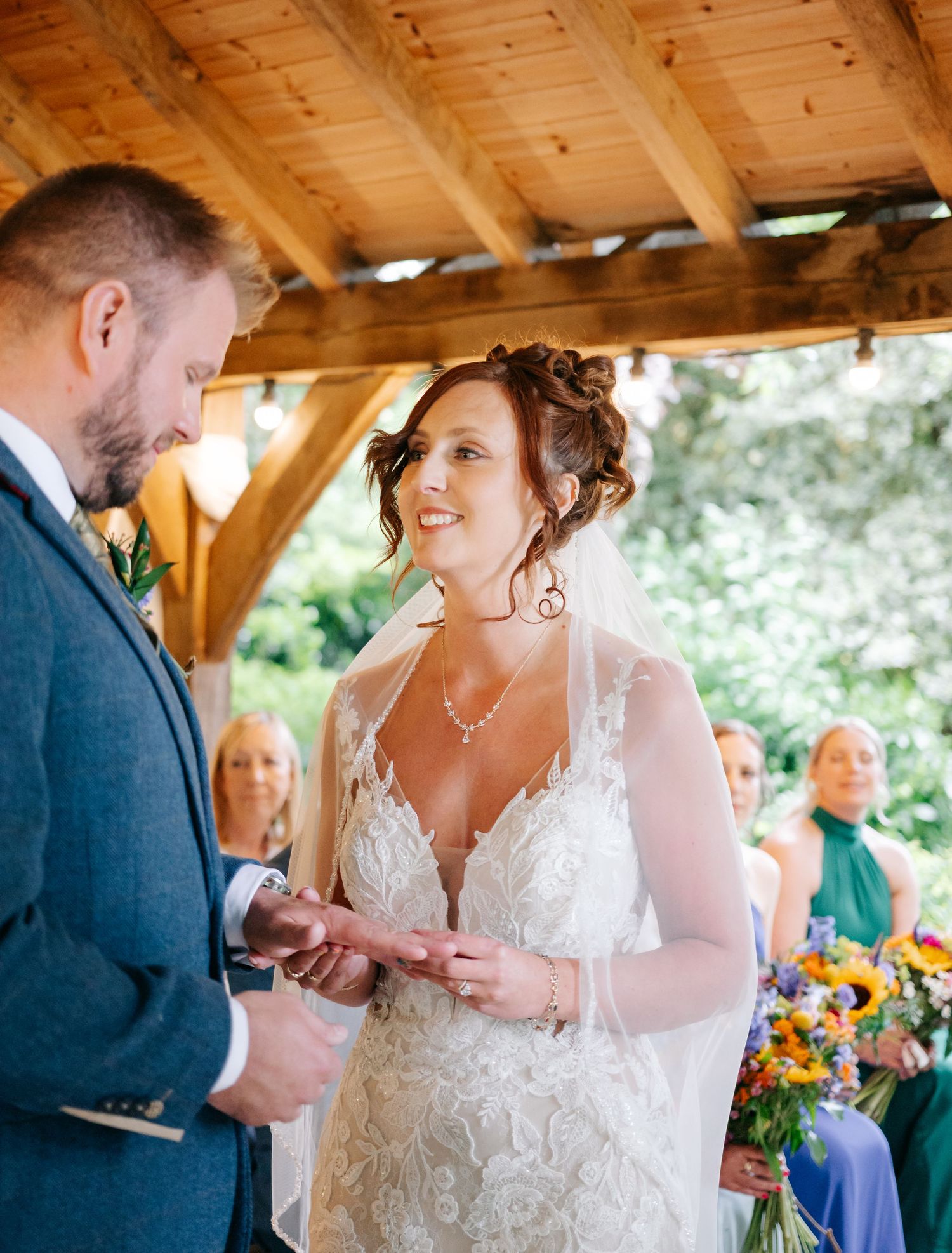 A wedding couple exchanges rings during their ceremony under a rustic wooden pergola.