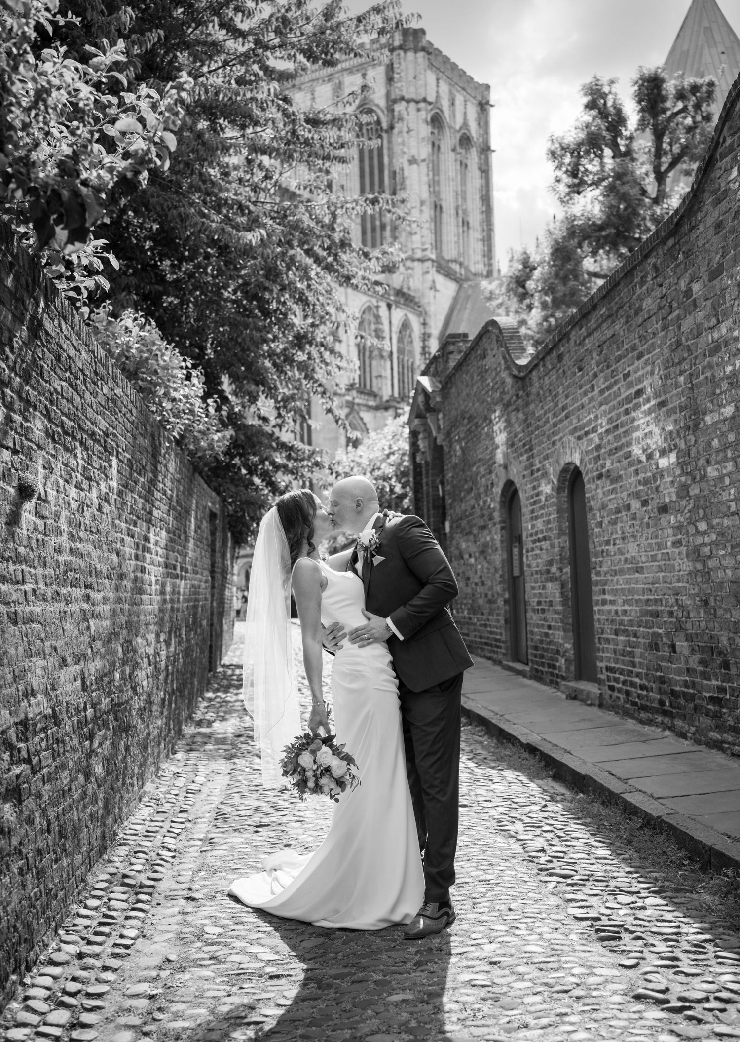 Black and white photo of a bride and groom kissing on a cobblestone alley with brick walls and a cathedral backdrop.