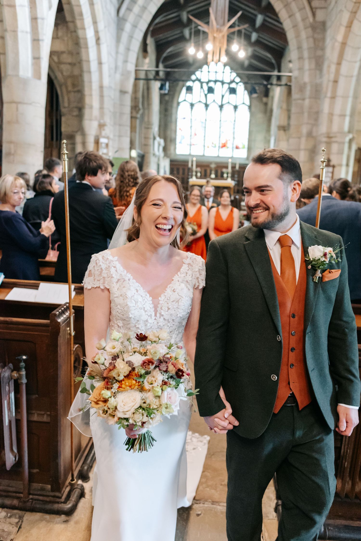 Smiling bride in a lace gown holding a bouquet walks hand in hand with her groom down the church aisle as guests look on.