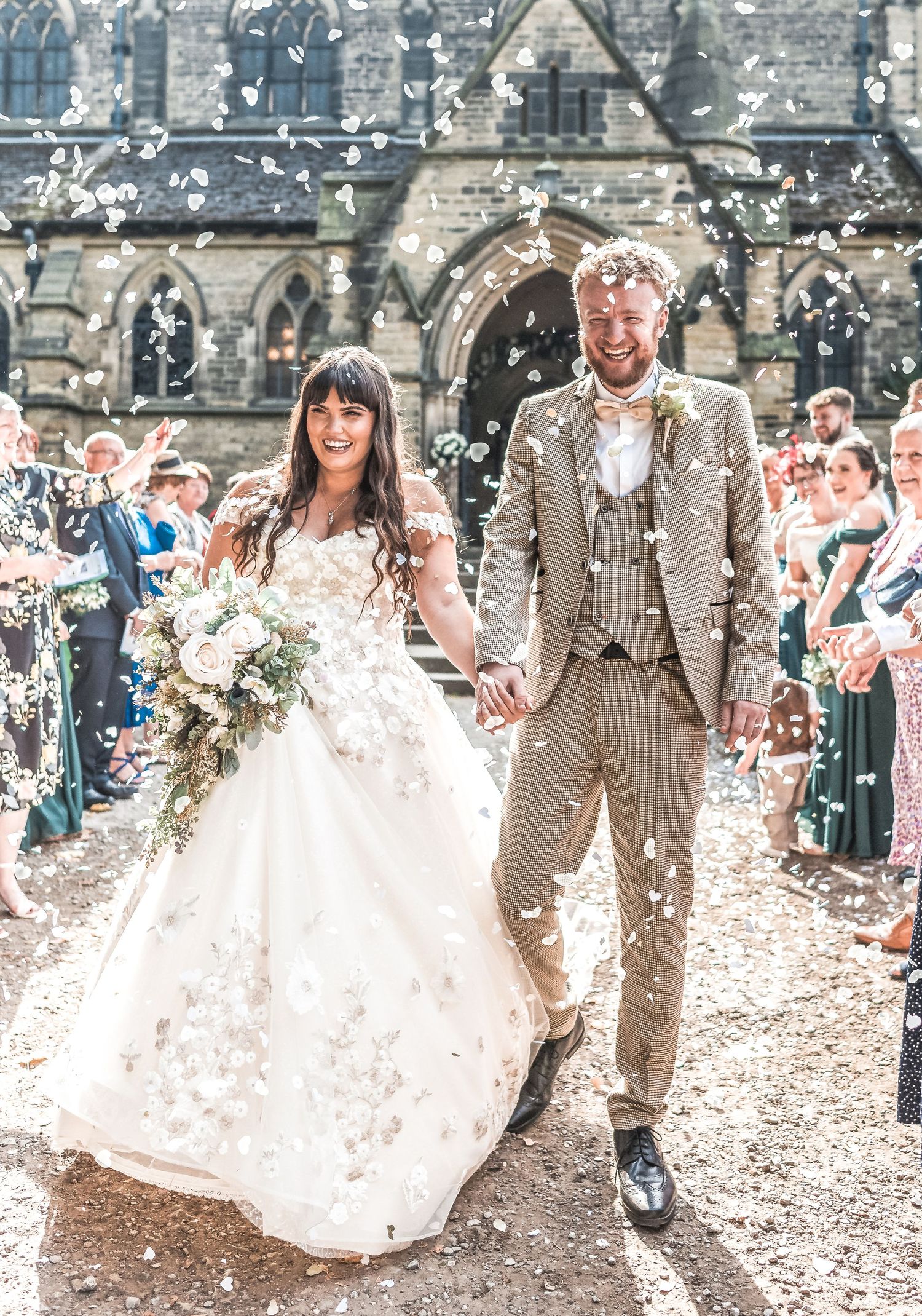 Wedding party walks through falling confetti outside a historic stone church while guests celebrate the joyous occasion.