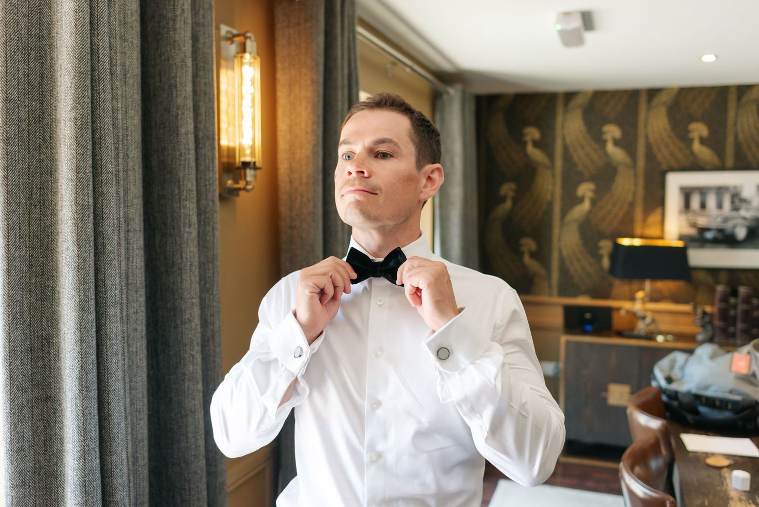 A person in a white dress shirt adjusts their bowtie while standing in an elegant room with ornate decor.