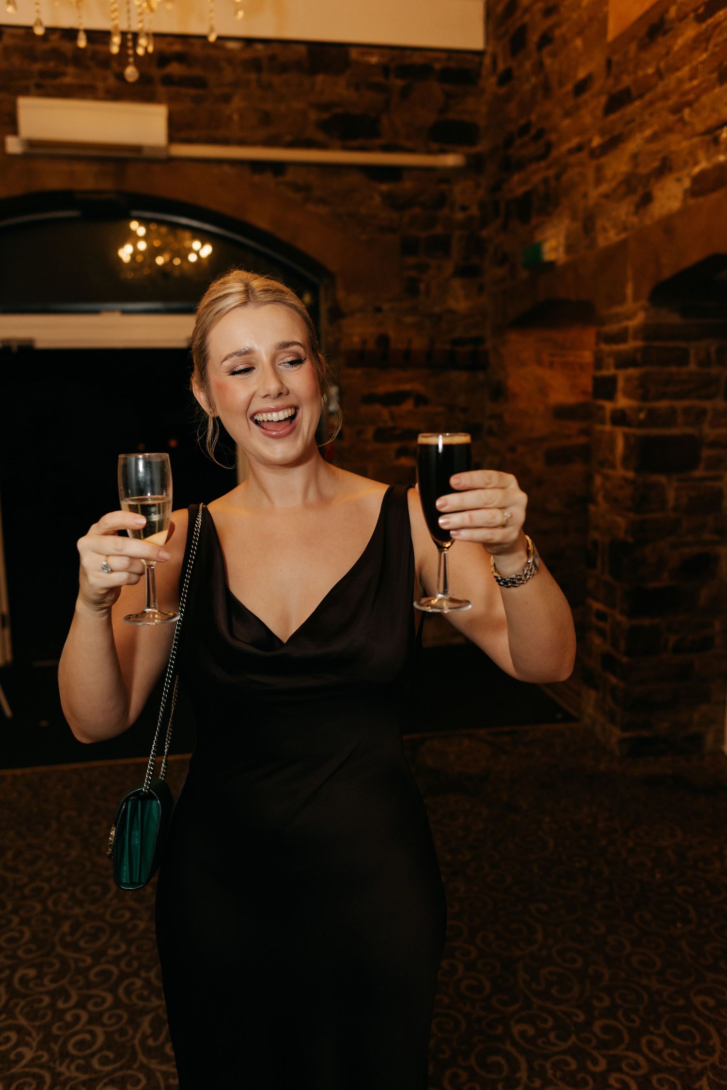 Person in black dress raises champagne glasses in celebration against brick wall backdrop.