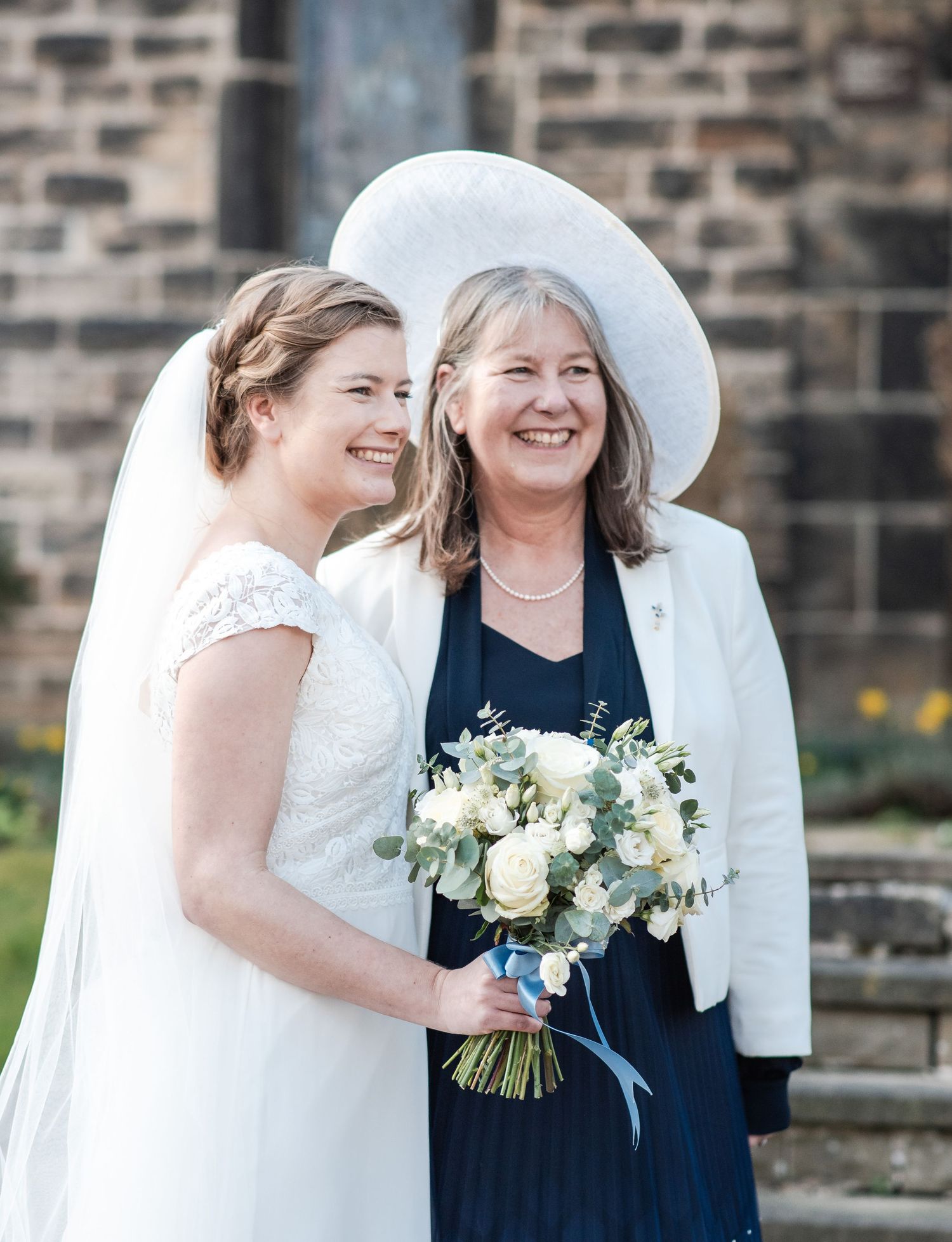 Wedding party members in white and navy share laughs while holding bouquets outside a stone church.