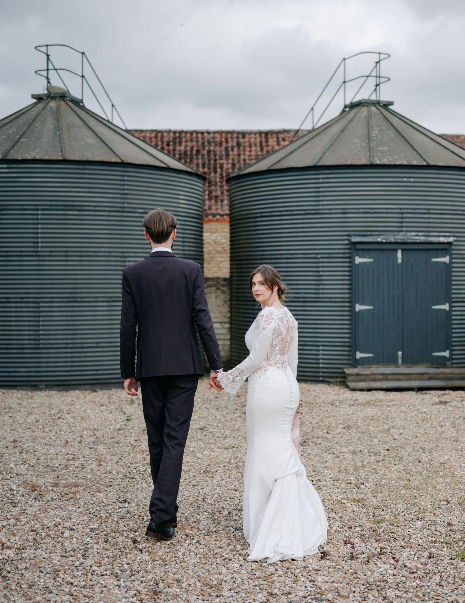 Couple walking near industrial green storage silos on gravel path.