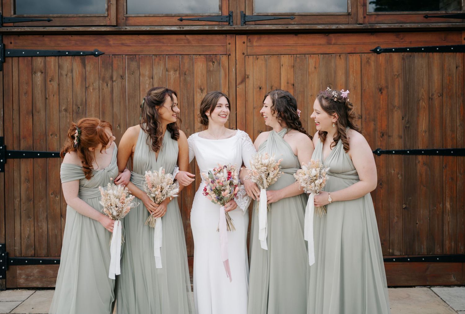 Bridal party in sage green dresses poses together against rustic wooden backdrop holding pink bouquets.