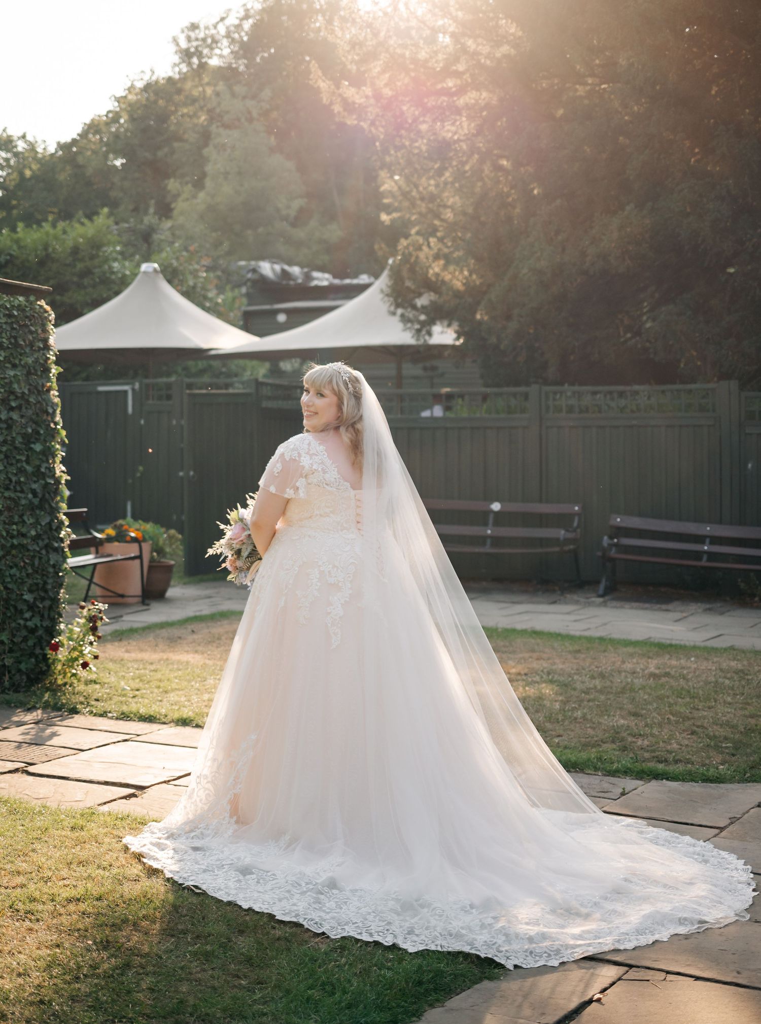 Stunning bride in lace ballgown with cathedral veil poses in golden sunlight at outdoor wedding venue.