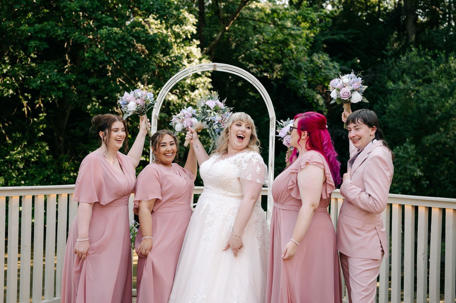 Bride in white gown laughs with bridesmaids in pink dresses under floral arch on wooden deck outdoors.