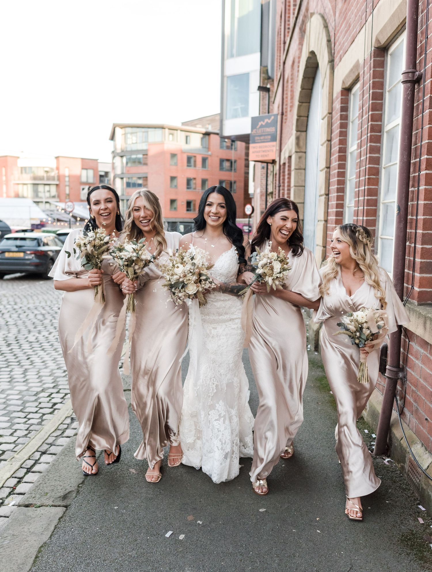 Bridesmaids in blush dresses walking together on cobblestone street holding bouquets.