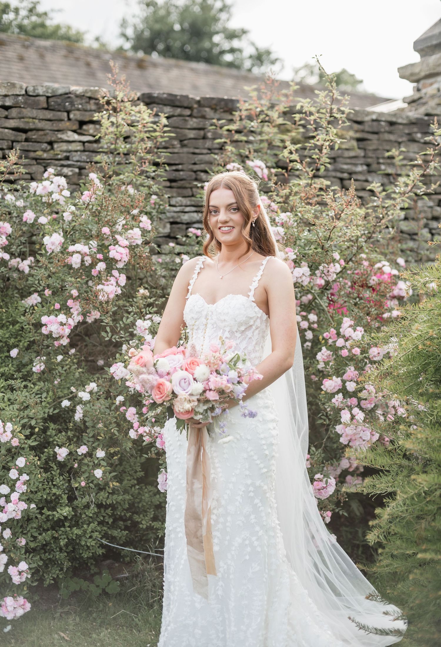 Elegant bride in lace dress poses against stone wall with pink blooming roses in garden setting.