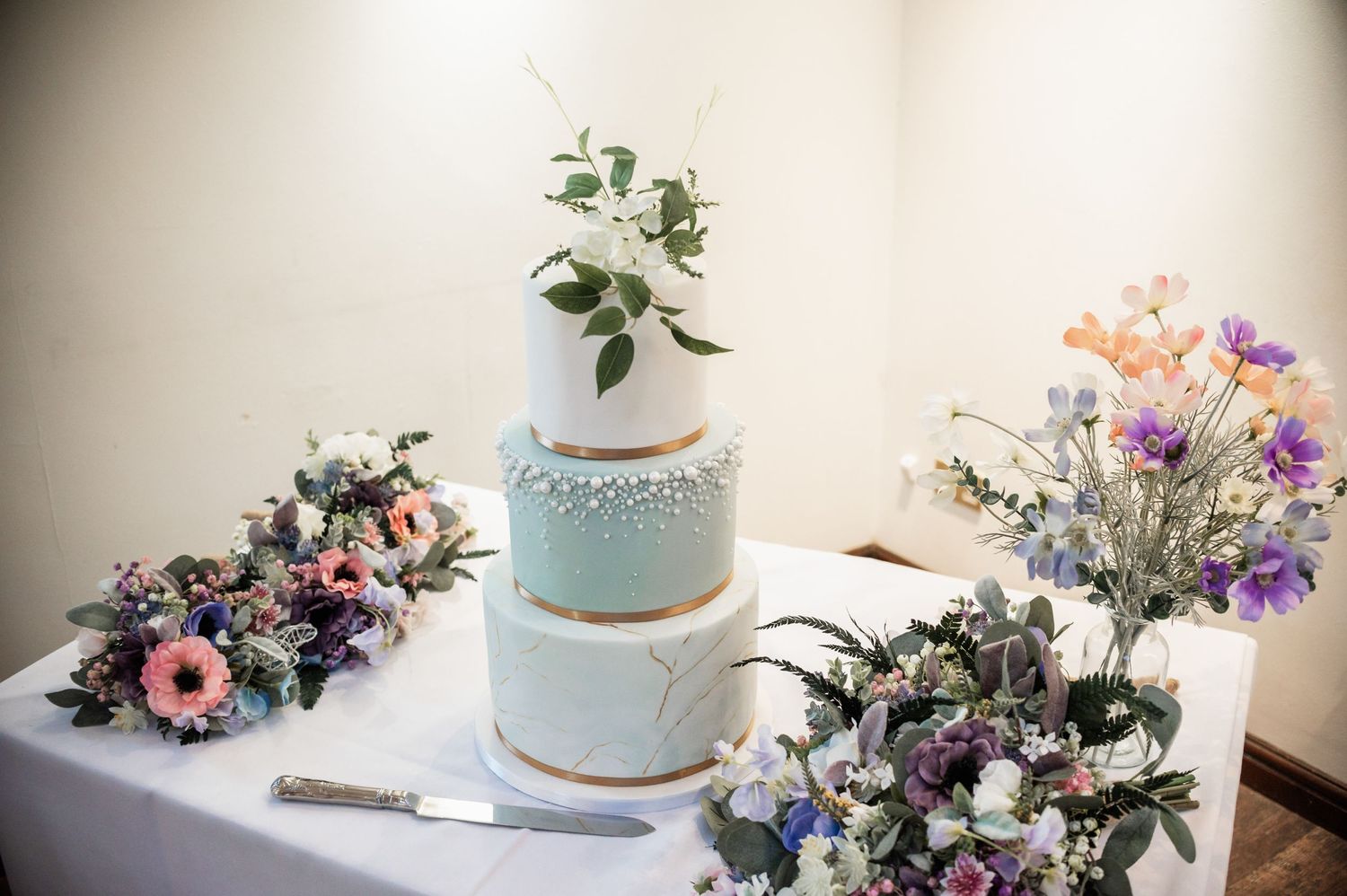 A pale blue wedding cake with floral decorations surrounded by colorful flower arrangements on a white table.