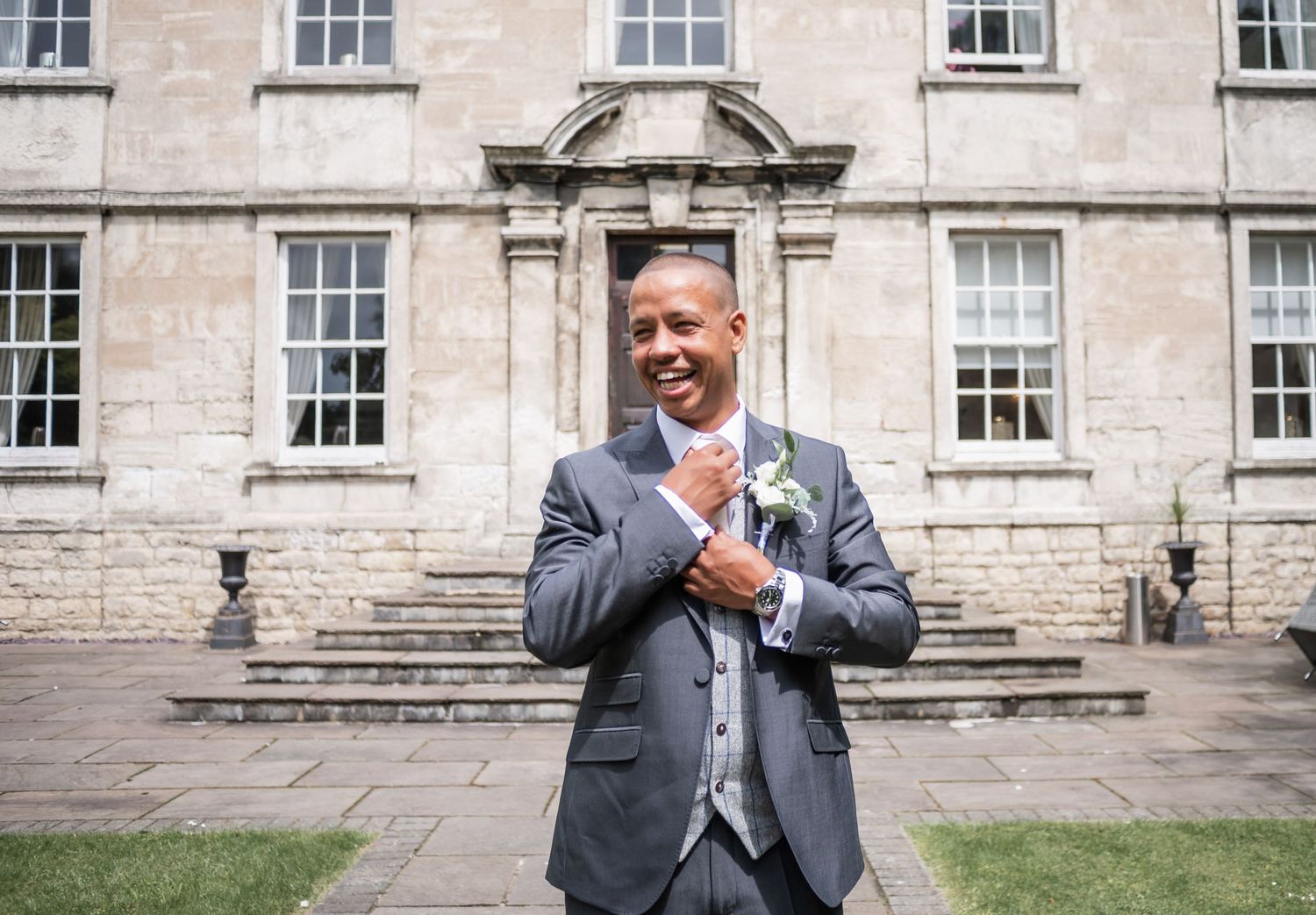 A person in a gray suit adjusts their tie while standing outside a grand stone building.