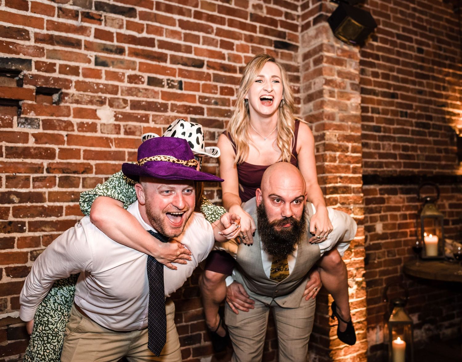 Wedding guests celebrate with energetic poses against a rustic brick wall backdrop during a reception.