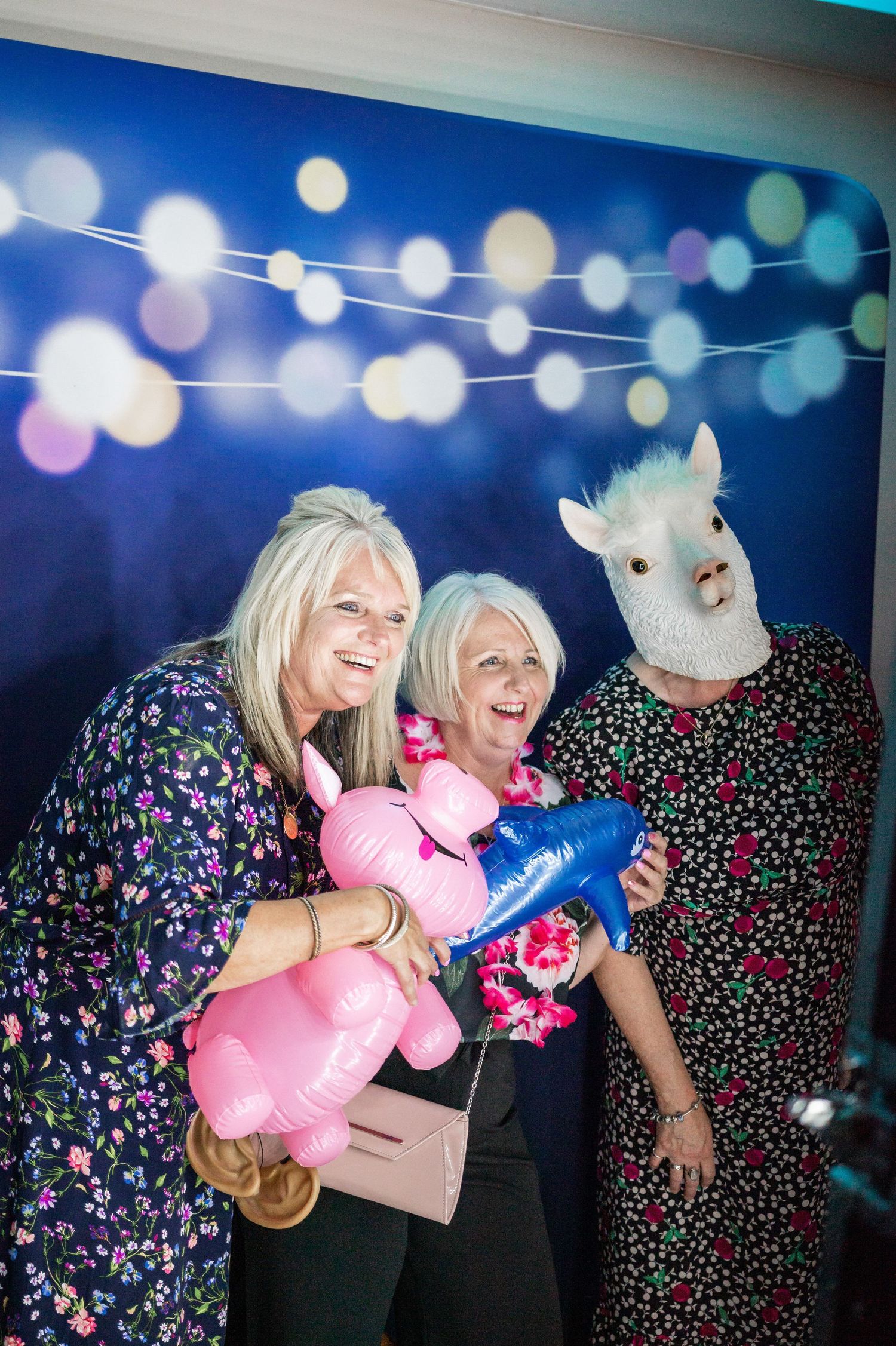 Party guests pose with balloons and a llama mascot against a backdrop with string lights.