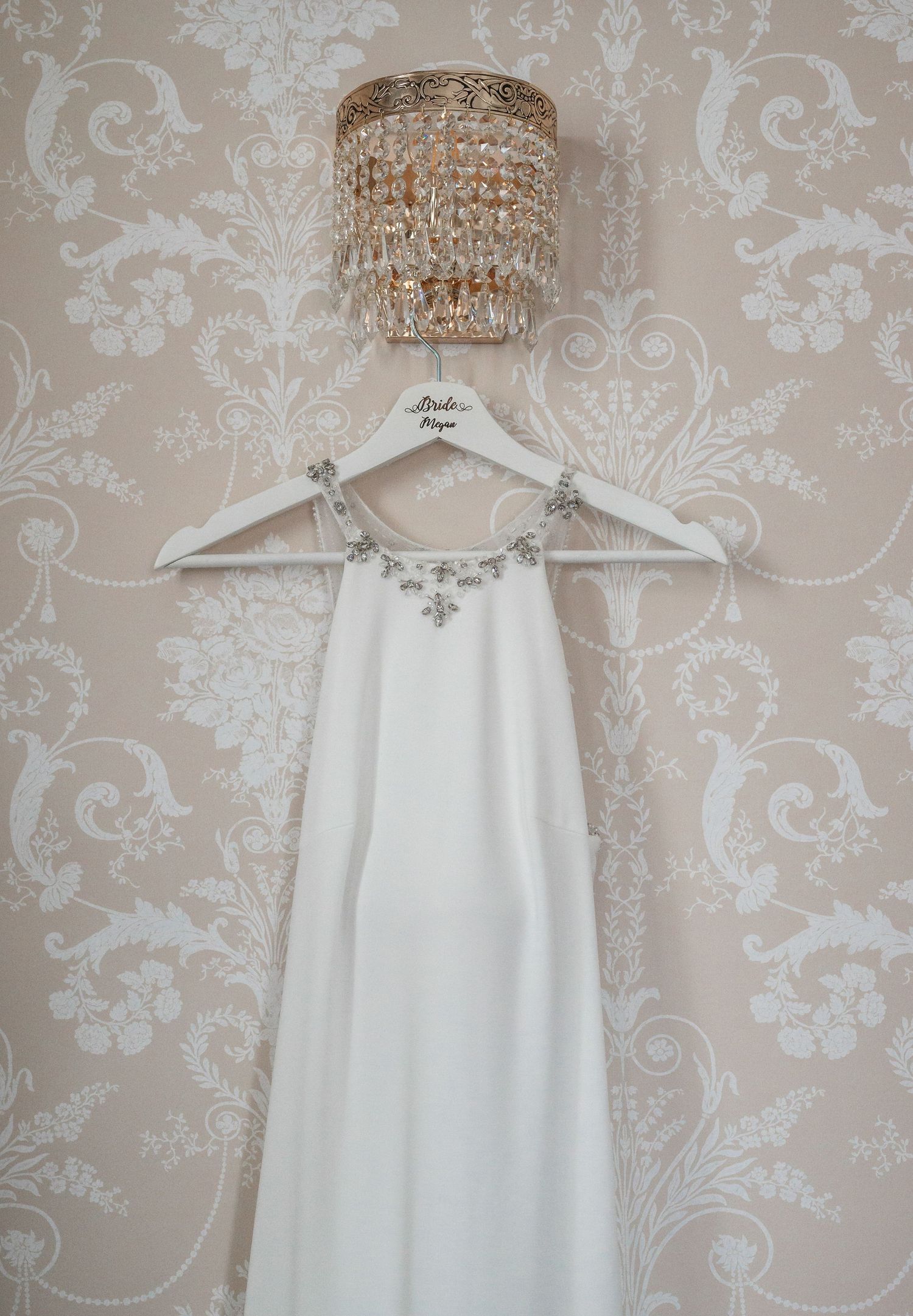 White wedding dress hanging under crystal chandelier against damask wallpaper.