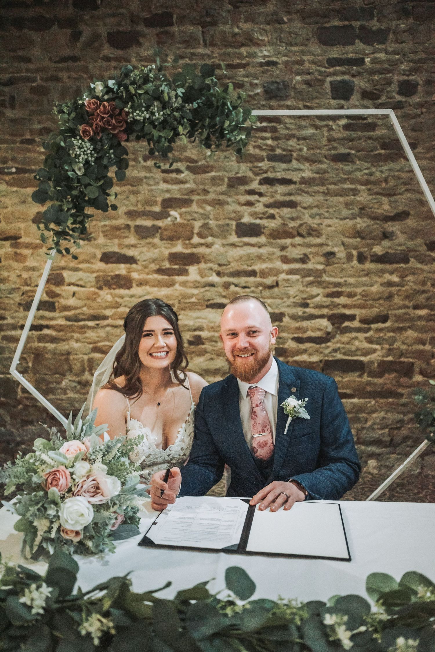 Couple signing wedding documents under a geometric arch with floral decorations.