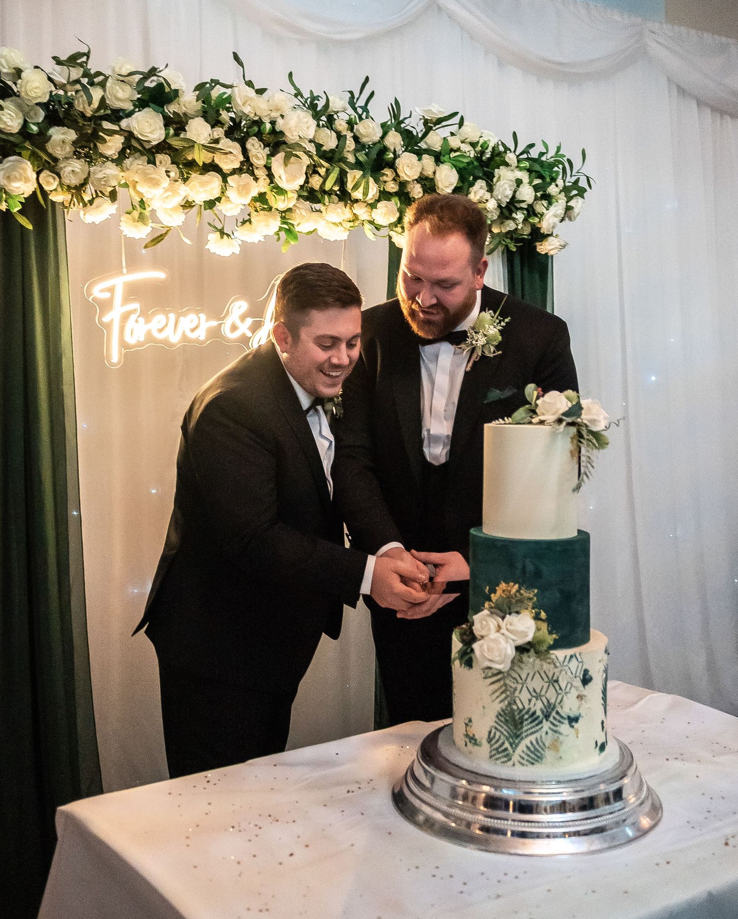 Newlyweds cutting elegant white and blue wedding cake under floral arch with neon sign.