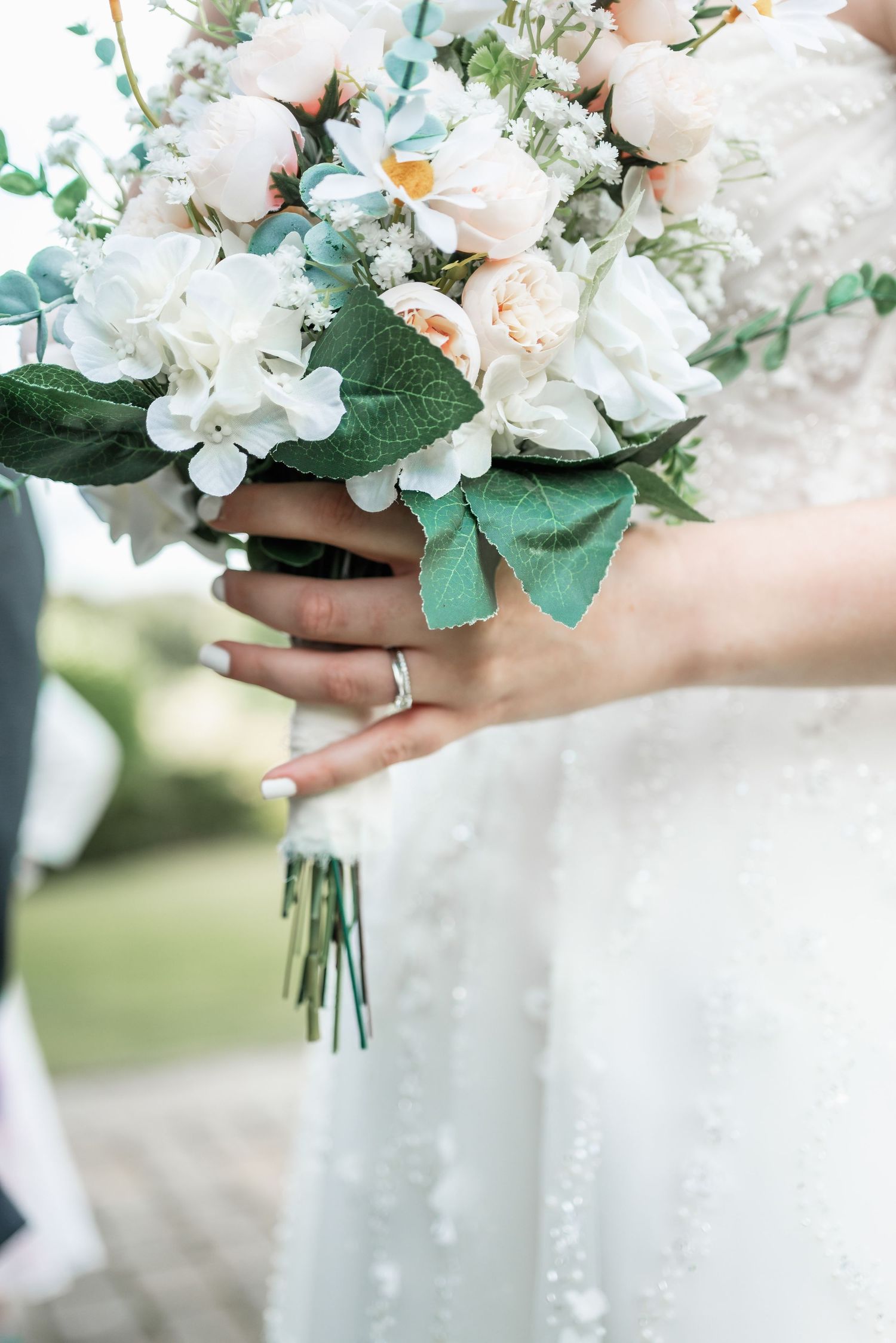 Delicate white and peach bridal bouquet with eucalyptus leaves held by hands showing wedding ring.