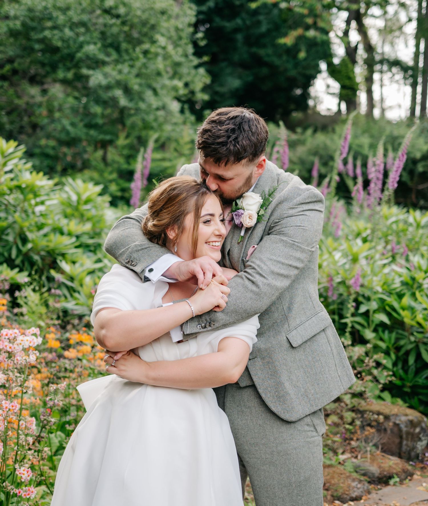 A couple shares an intimate embrace in a lush garden filled with pink and yellow blooms and tall evergreens.