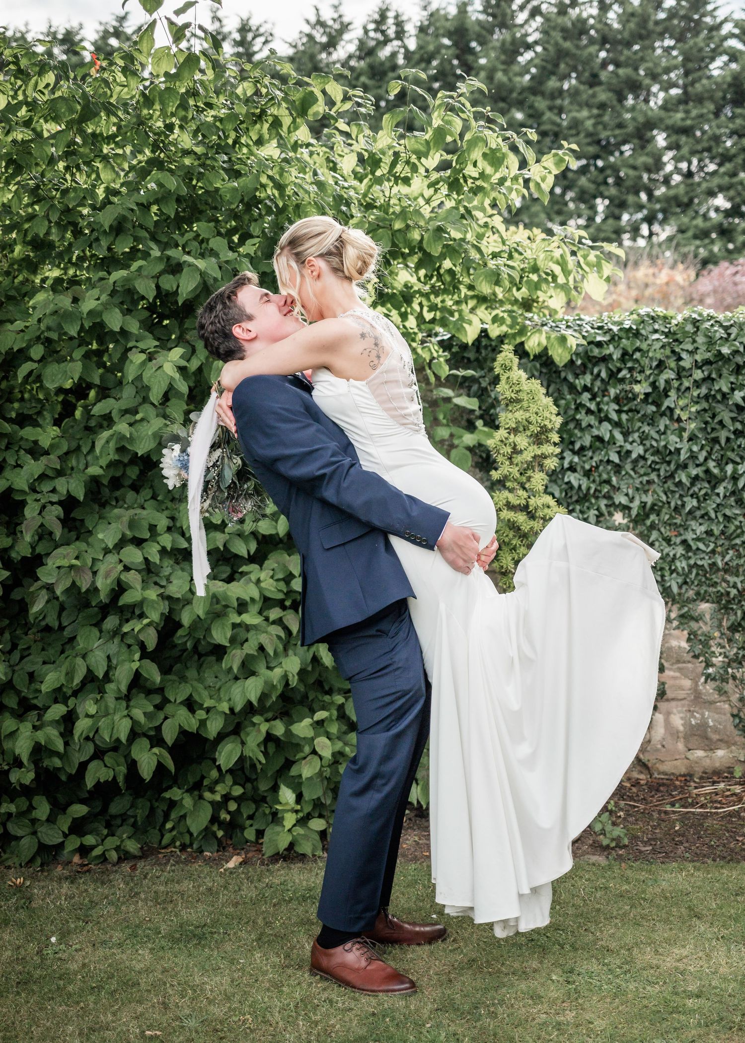 Playful wedding portrait with groom lifting bride in garden setting surrounded by greenery.
