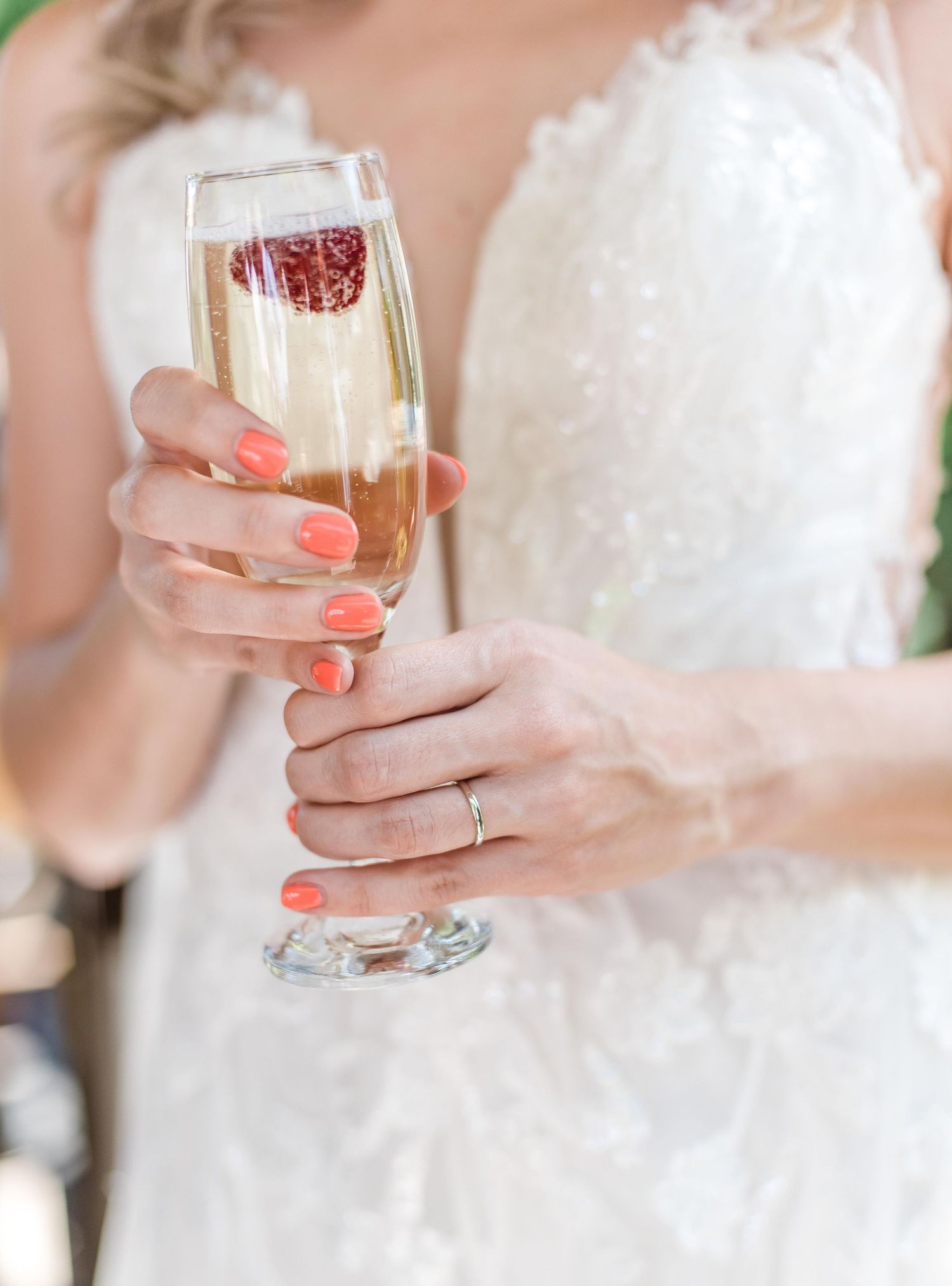Elegant hand with coral nails holding champagne flute against white wedding dress.