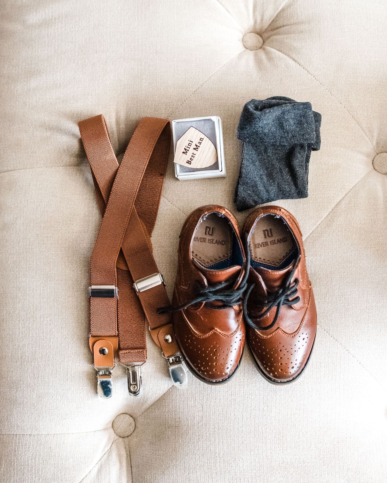 Brown leather suspenders and dress shoes arranged on cream tufted surface.