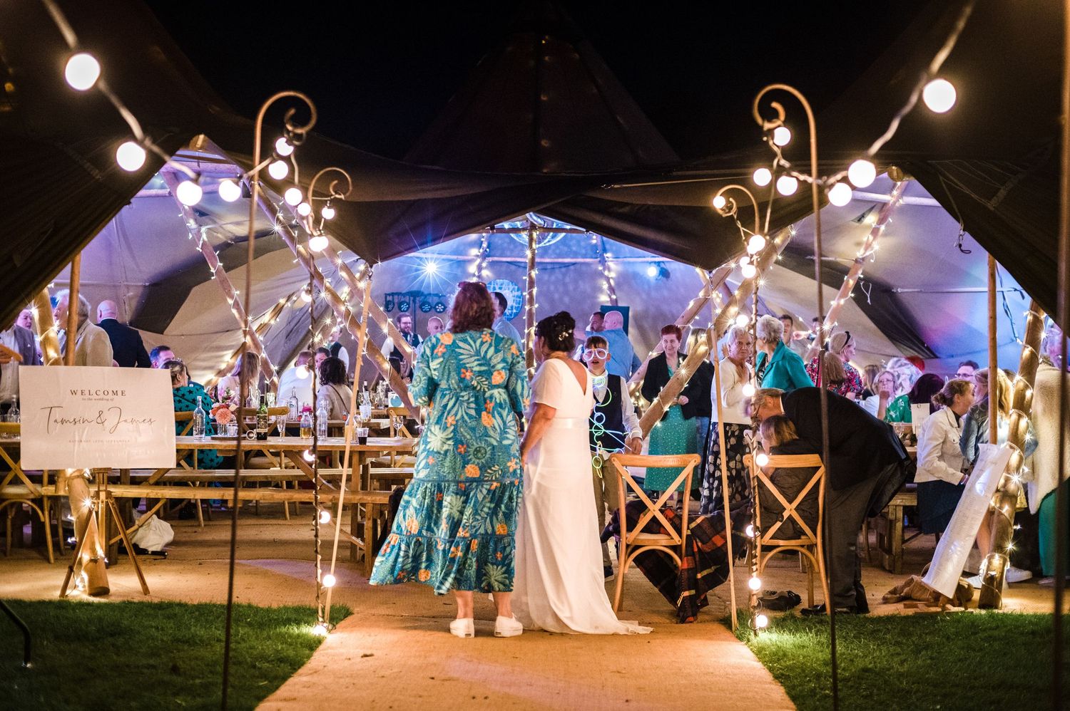Wedding reception under a lit tent with guests dancing on a wooden pathway.