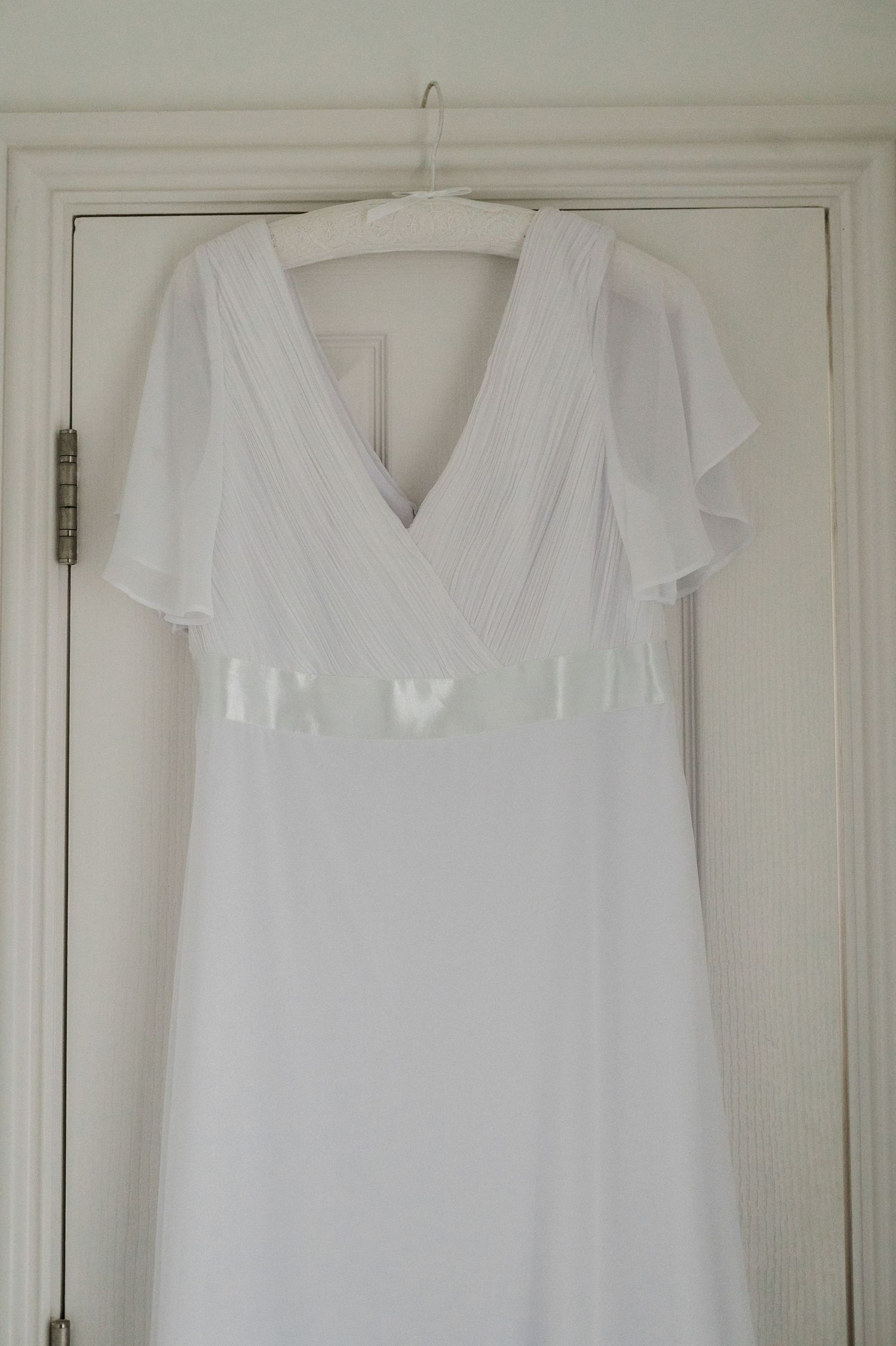 A white wedding dress with flutter sleeves and empire waist hanging on a white door.