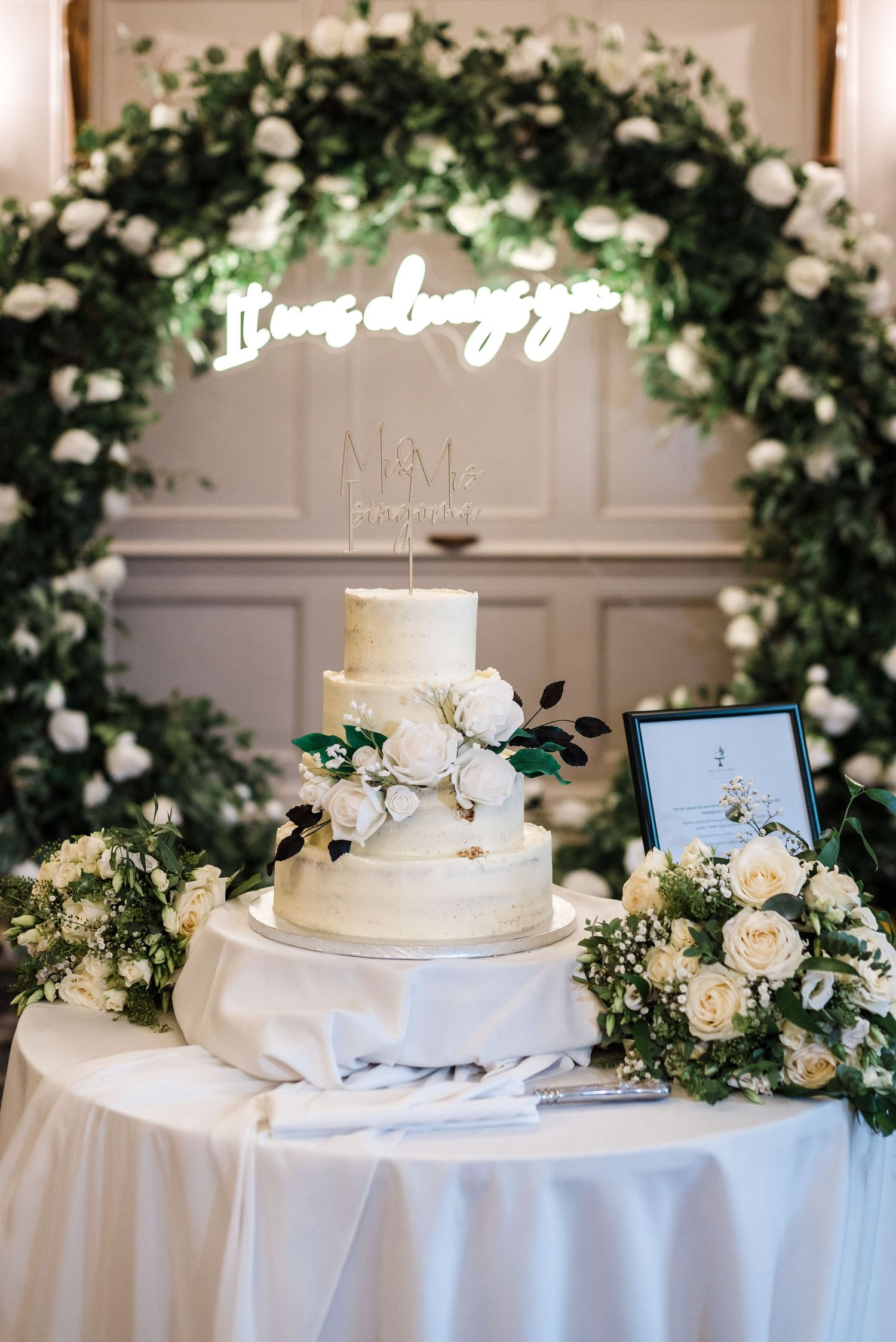 Elegant white wedding cake displayed on table with floral arch and neon sign decor.