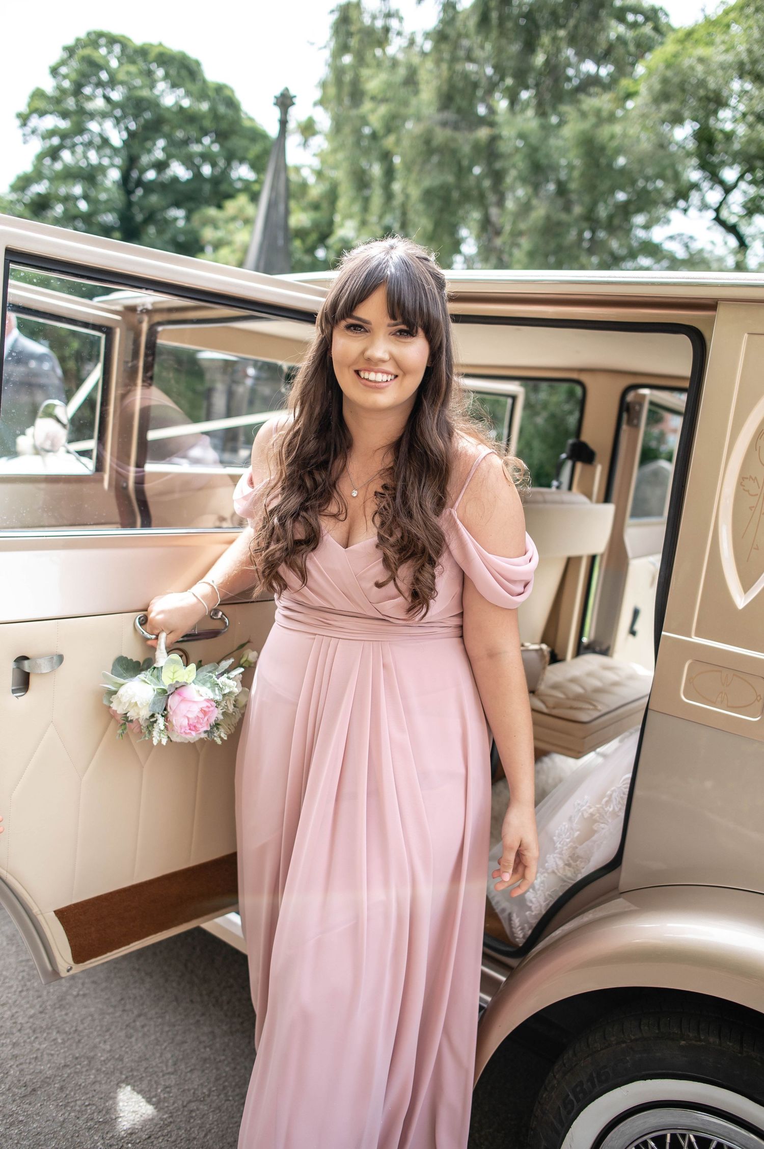 Bridesmaid in pink dress holding bouquet next to vintage cream-colored wedding car.