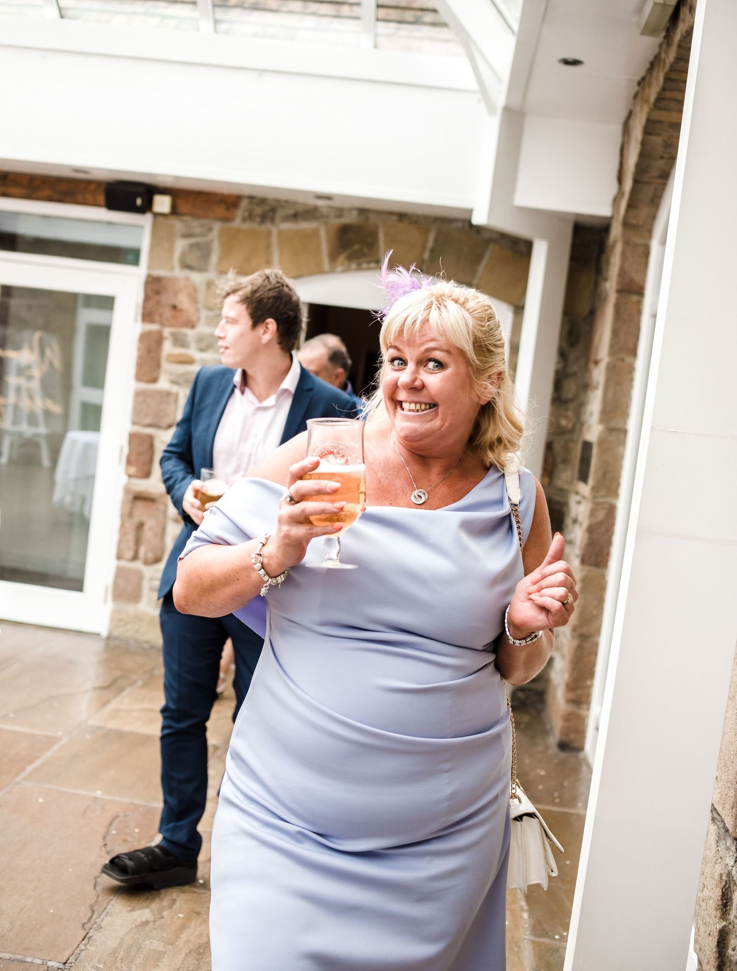 Wedding guest in grey dress holds champagne glass while smiling at outdoor venue.