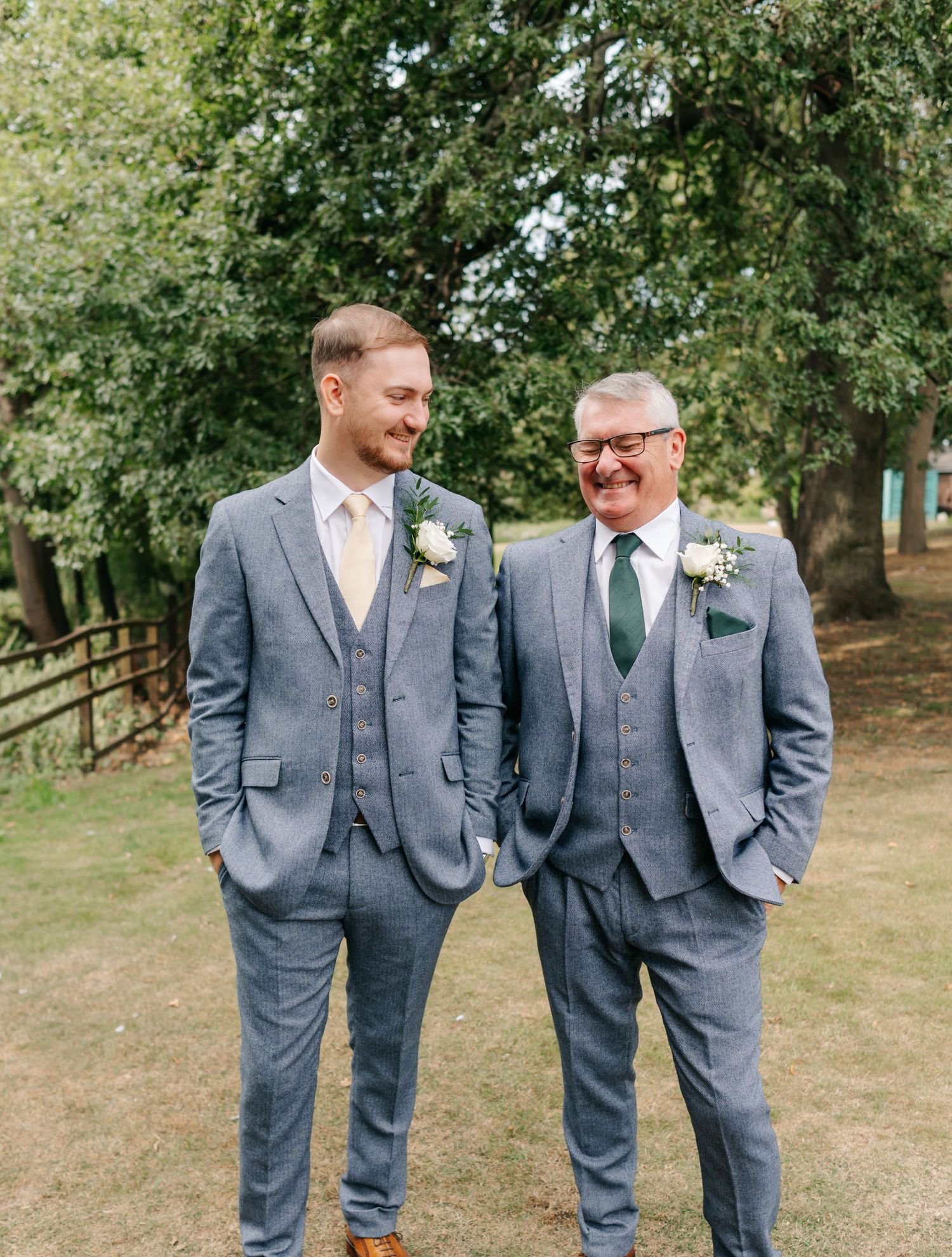 Two people wearing matching gray three-piece suits with boutonnieres stand together outdoors.