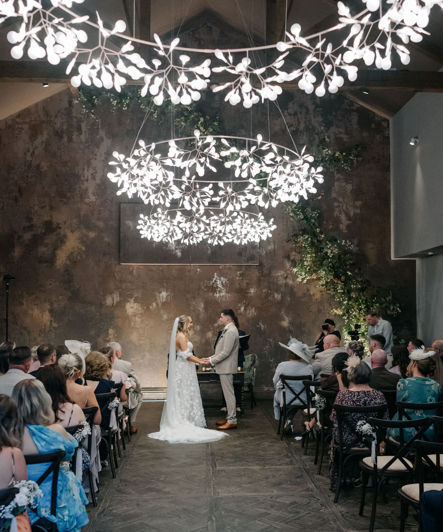 Wedding ceremony setup in a rustic venue with white floral ceiling decorations and dark wooden chairs.