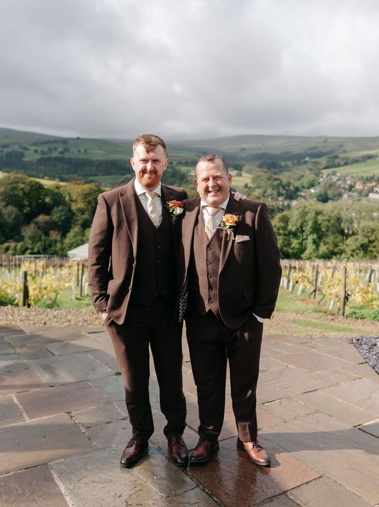 Groomsmen in brown suits pose together on a stone patio with rolling hills in the background.