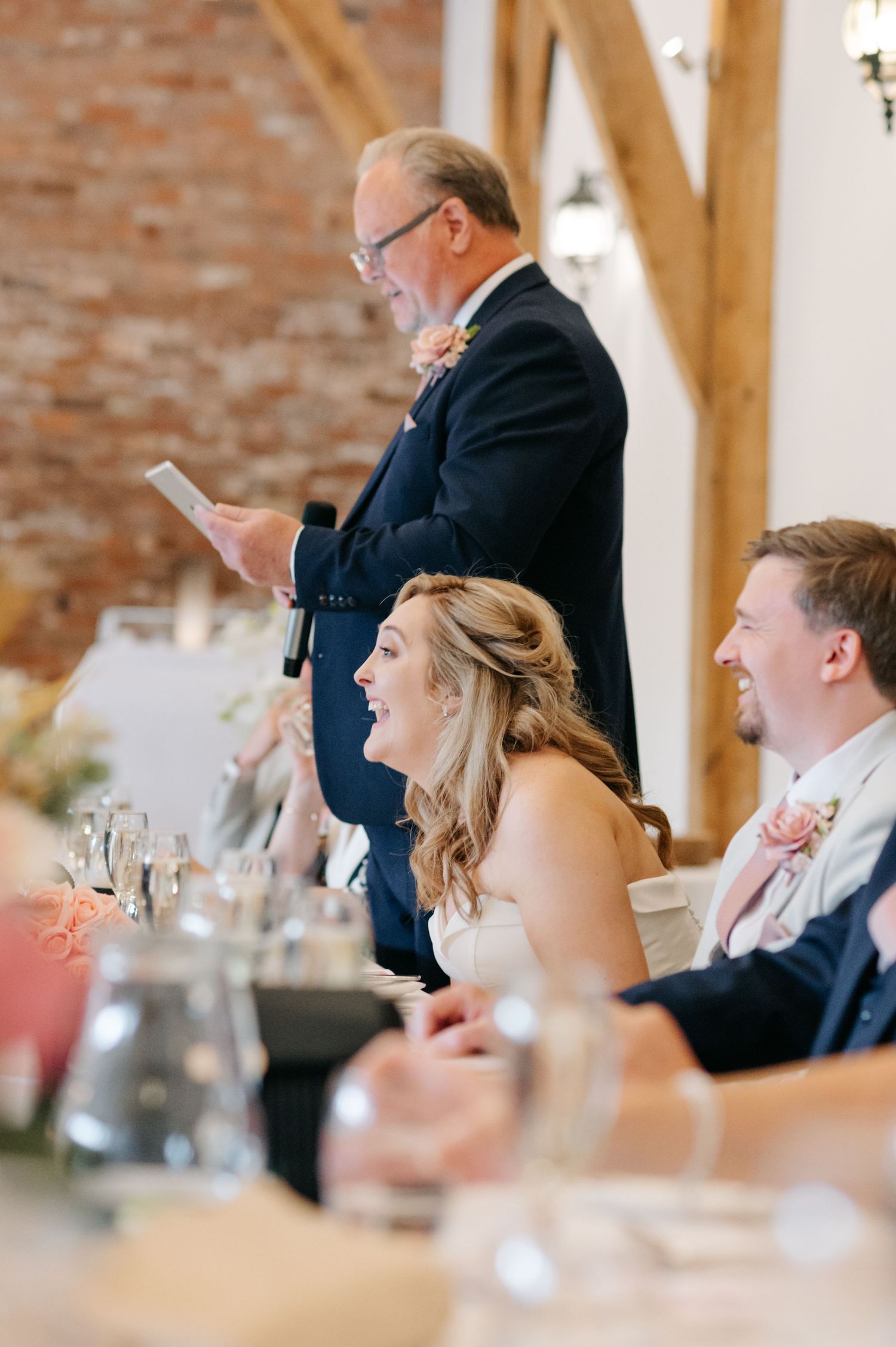 Wedding reception guests seated at long tables in rustic venue with exposed brick walls and wooden beams.