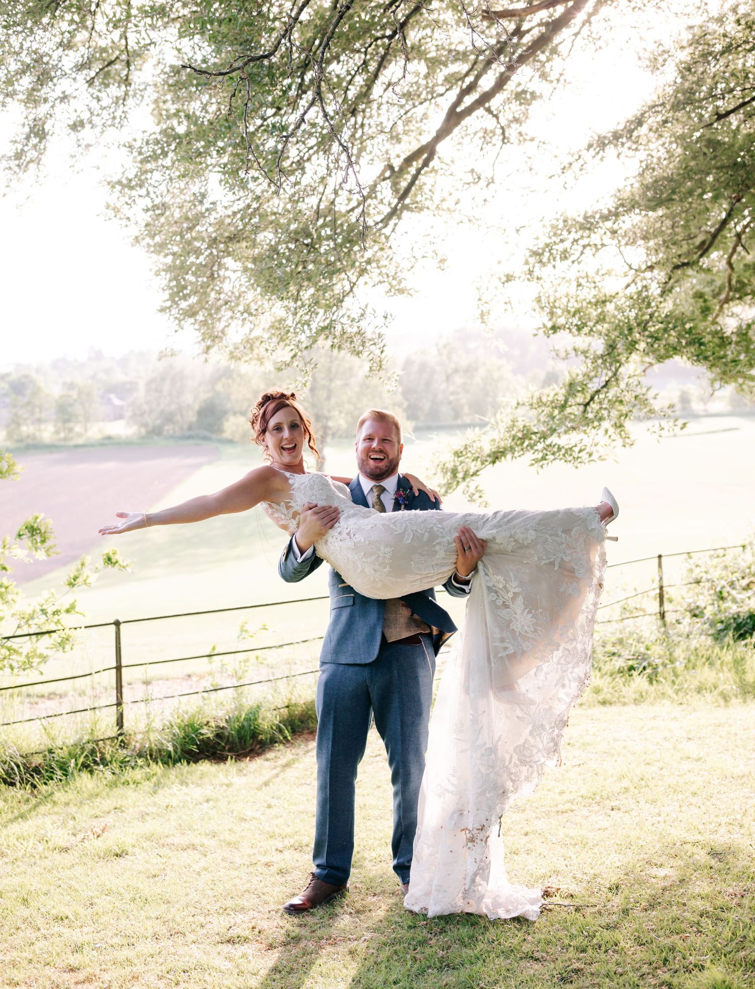 Groom in blue suit carries bride in white lace dress against scenic lake backdrop.