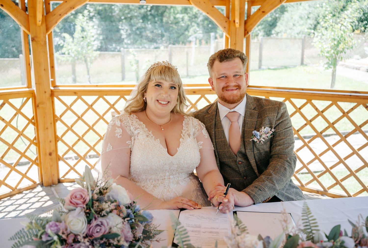 A happy bride and groom pose together signing their wedding register under a wooden gazebo.