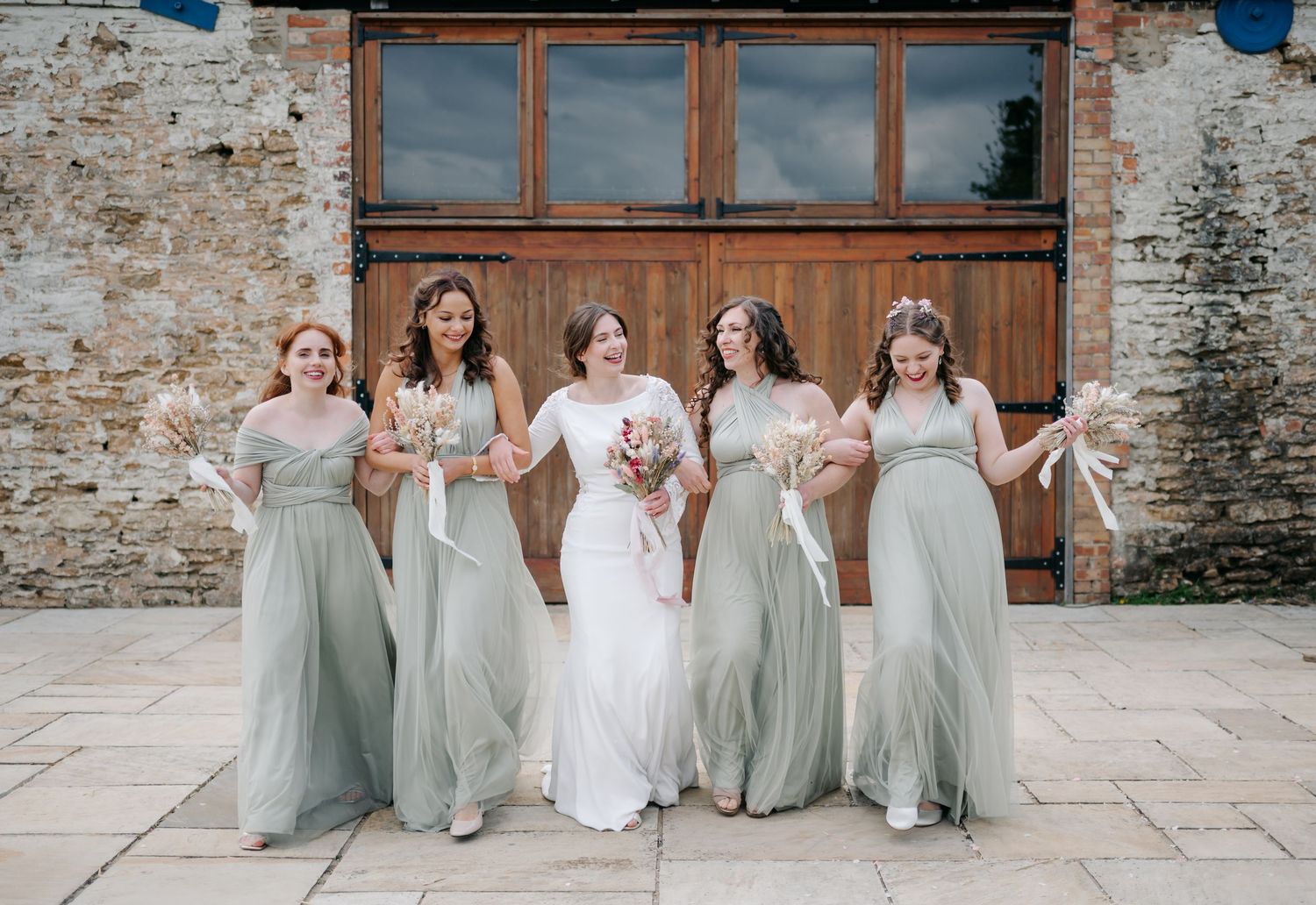Wedding party in sage green bridesmaid dresses seated against a rustic stone wall.