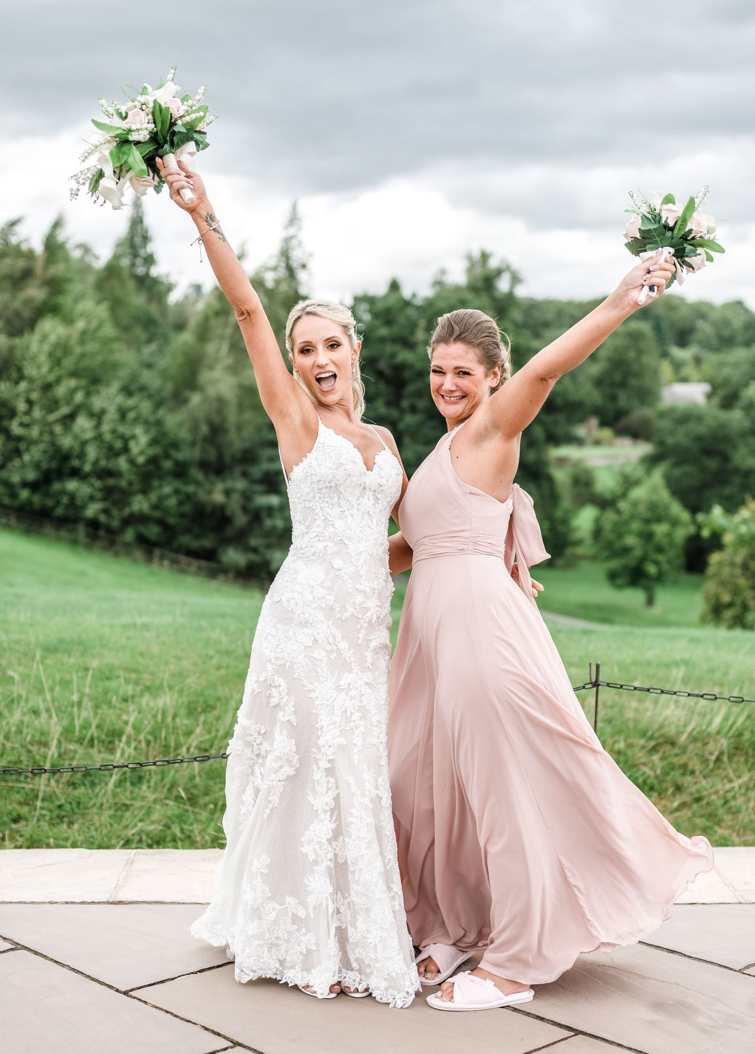 Bride and bridesmaid celebrate joyfully with raised bouquets on outdoor terrace.