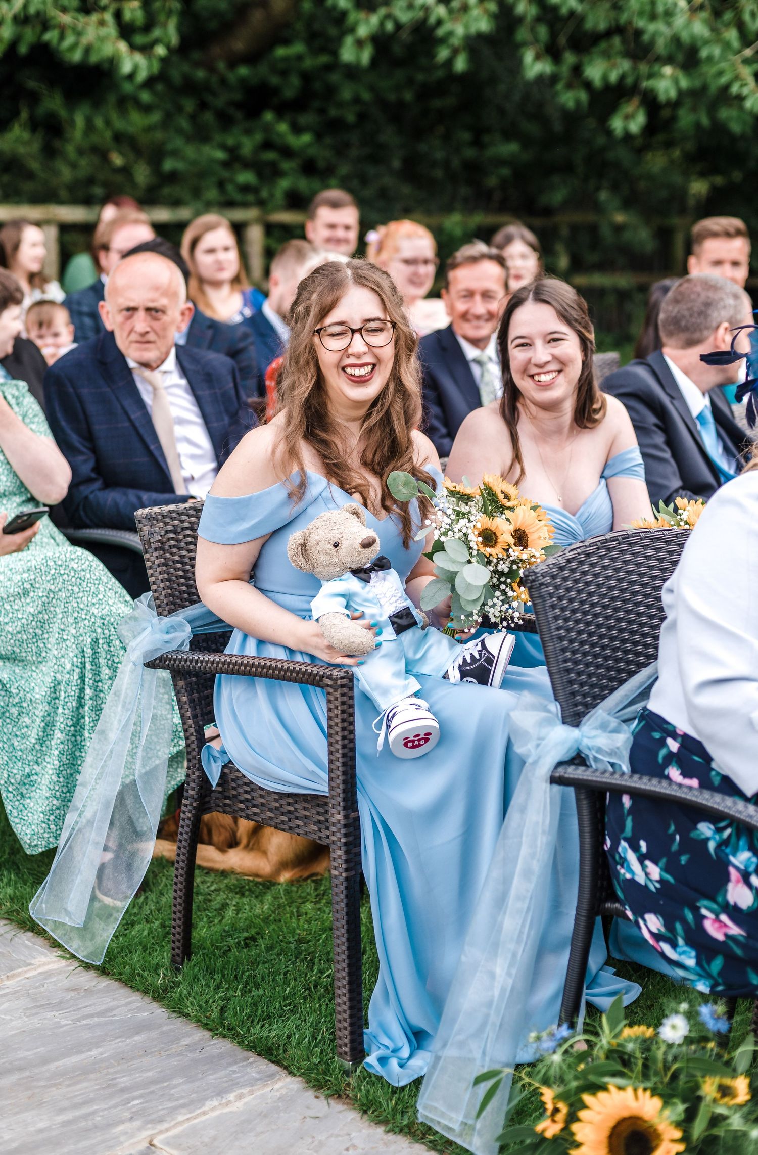 Wedding guests in blue dresses smile while holding sunflowers and a small plush toy during outdoor ceremony.