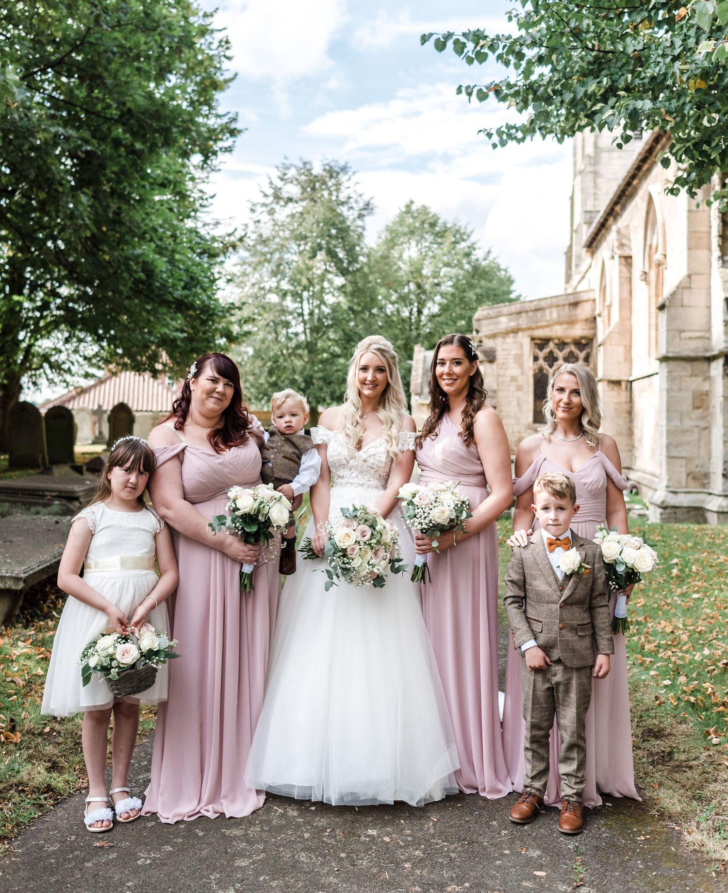 Wedding party in dusty pink bridesmaid dresses poses outside historic church with white bridal bouquets.