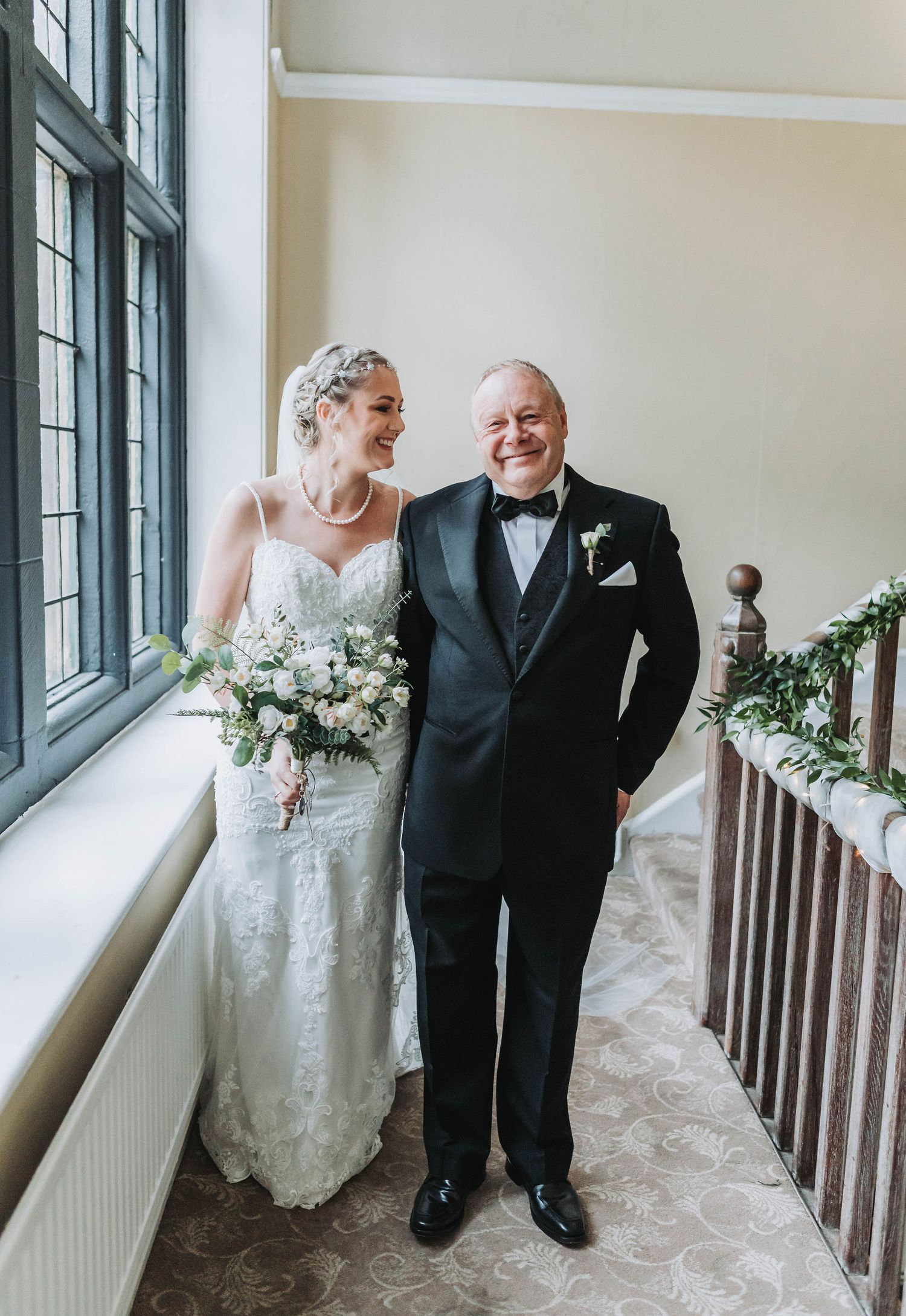 A bride in lace gown with bouquet poses by a window on her wedding day.