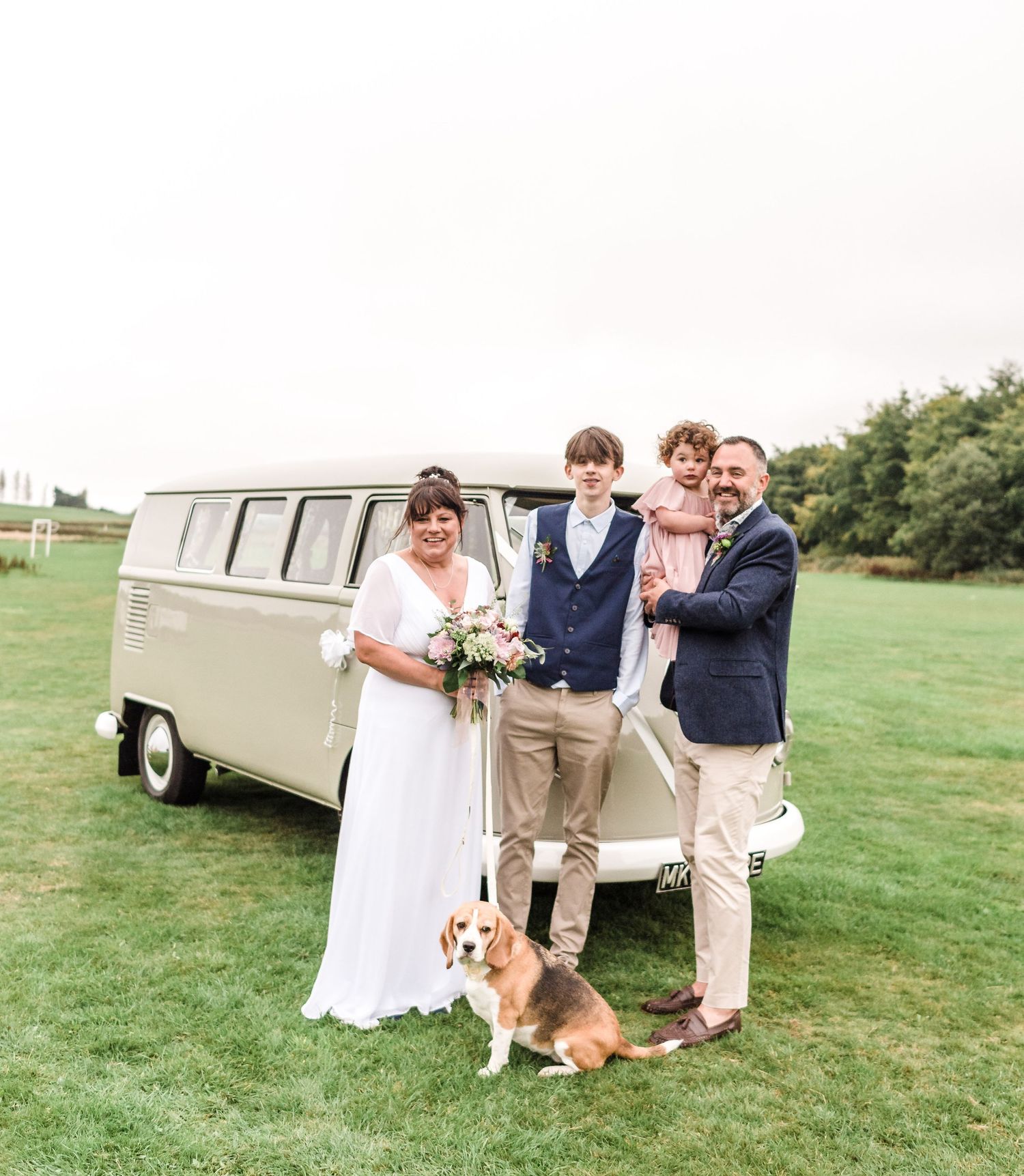 A wedding party poses with a vintage VW bus and corgi on a green lawn.